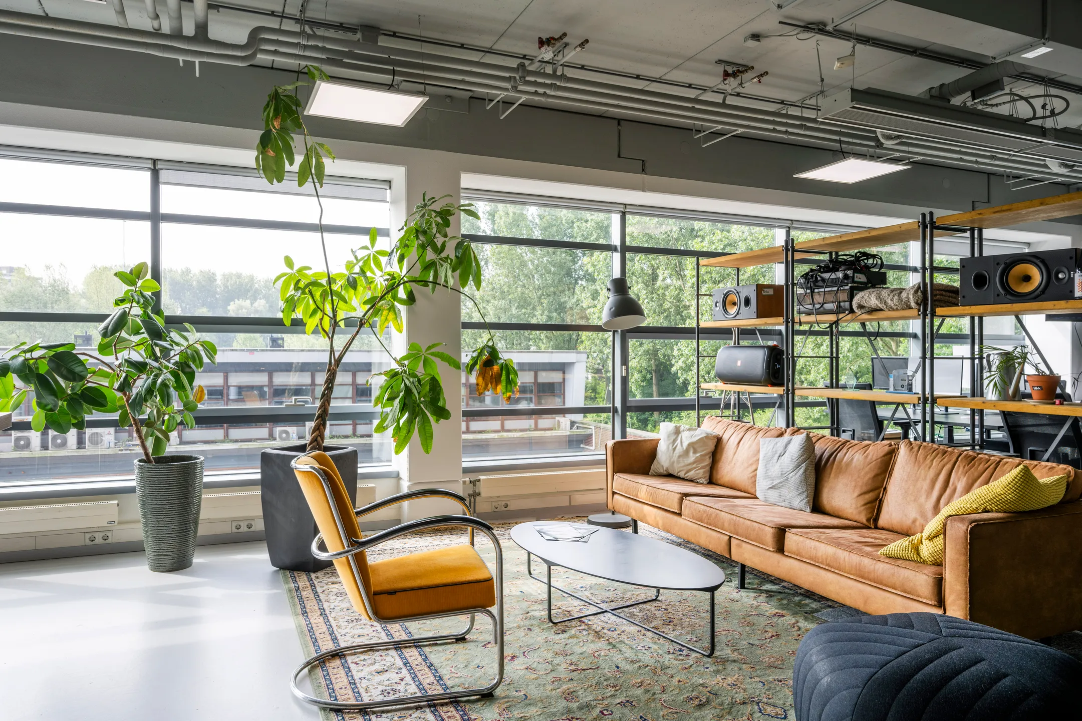 Een moderne woonkamer met grote ramen, een leren bank, een geelgestoffeerde stoel en veel planten.