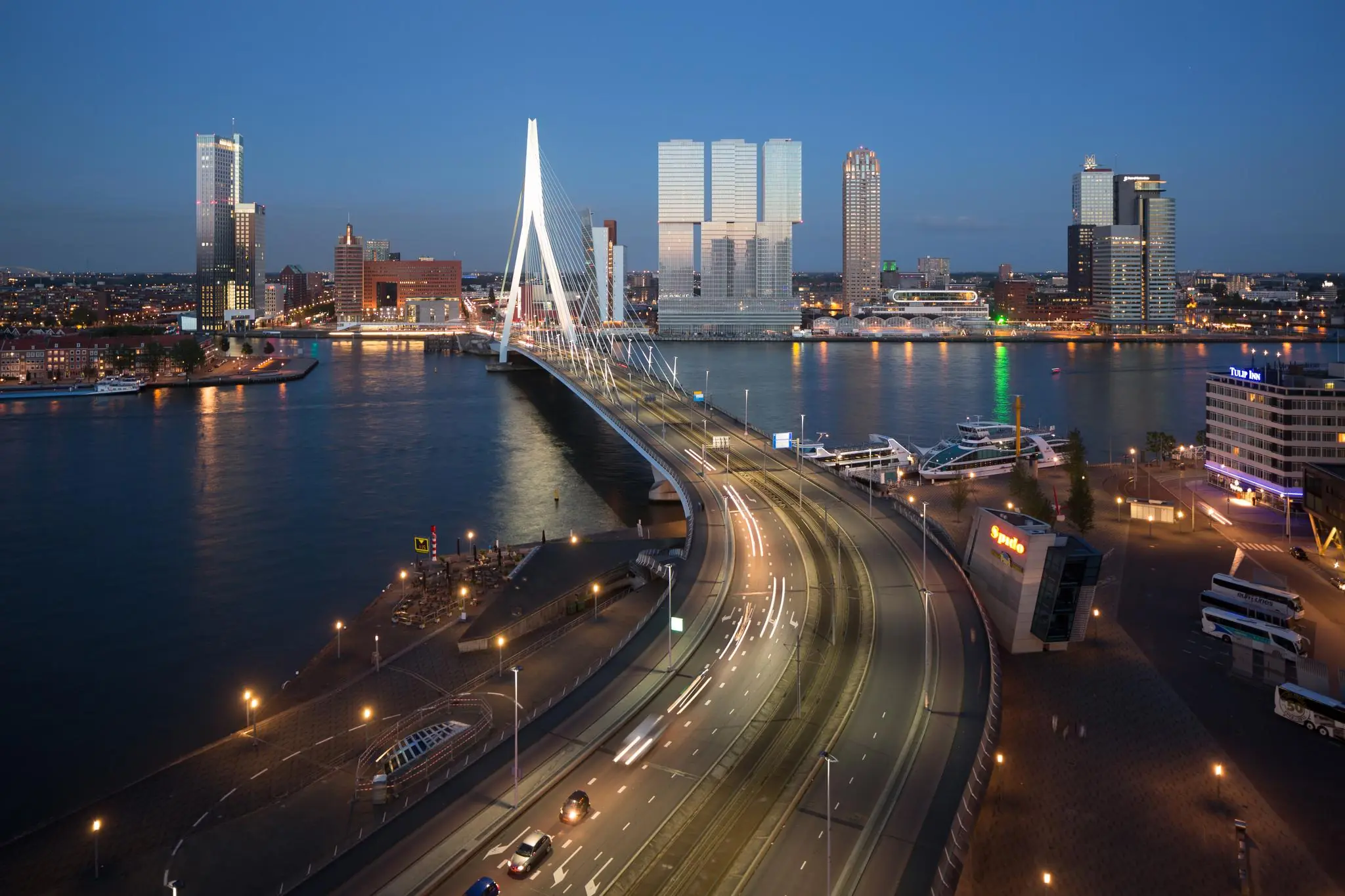 Avondzicht op de Wilhelminakade in Rotterdam met de verlichte Erasmusbrug en moderne hoogbouw op de achtergrond.