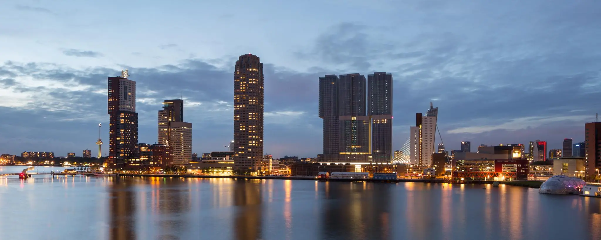 Avondzicht op de skyline van de Wilhelminakade in Rotterdam met verlichte hoogbouw en weerspiegeling in het water.