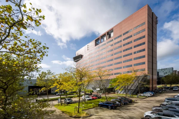 Groot modern gebouw met parkeerplaats en bomen op de voorgrond onder een blauwe hemel.