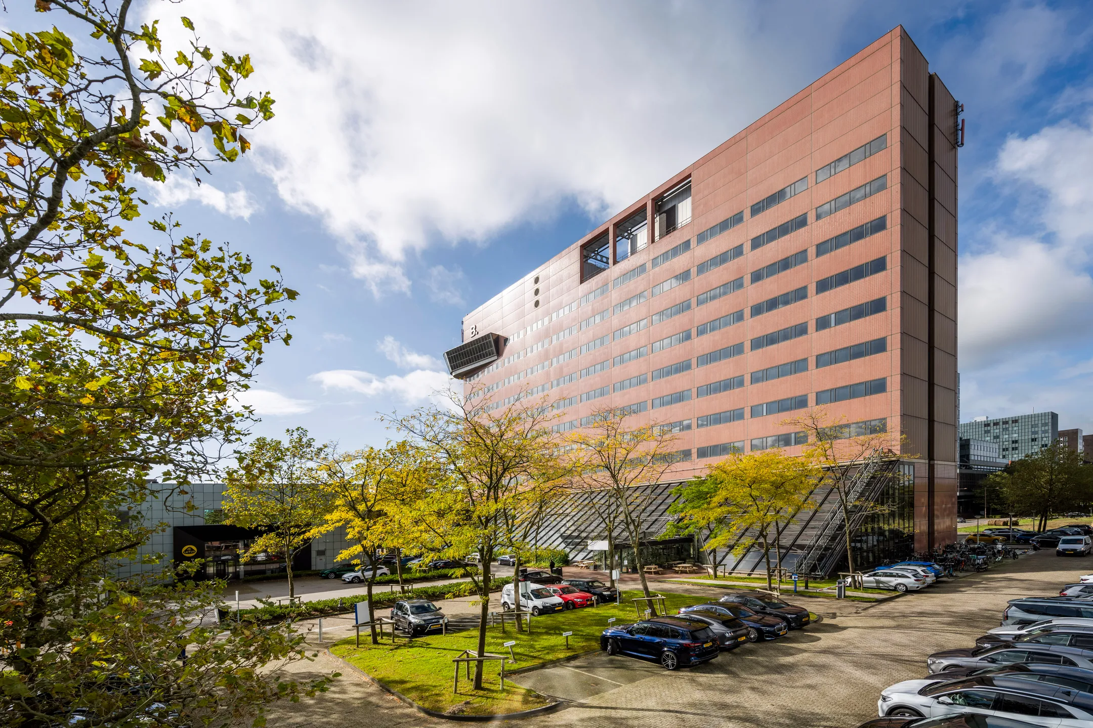 Groot modern gebouw met parkeerplaats en bomen op de voorgrond onder een blauwe hemel.