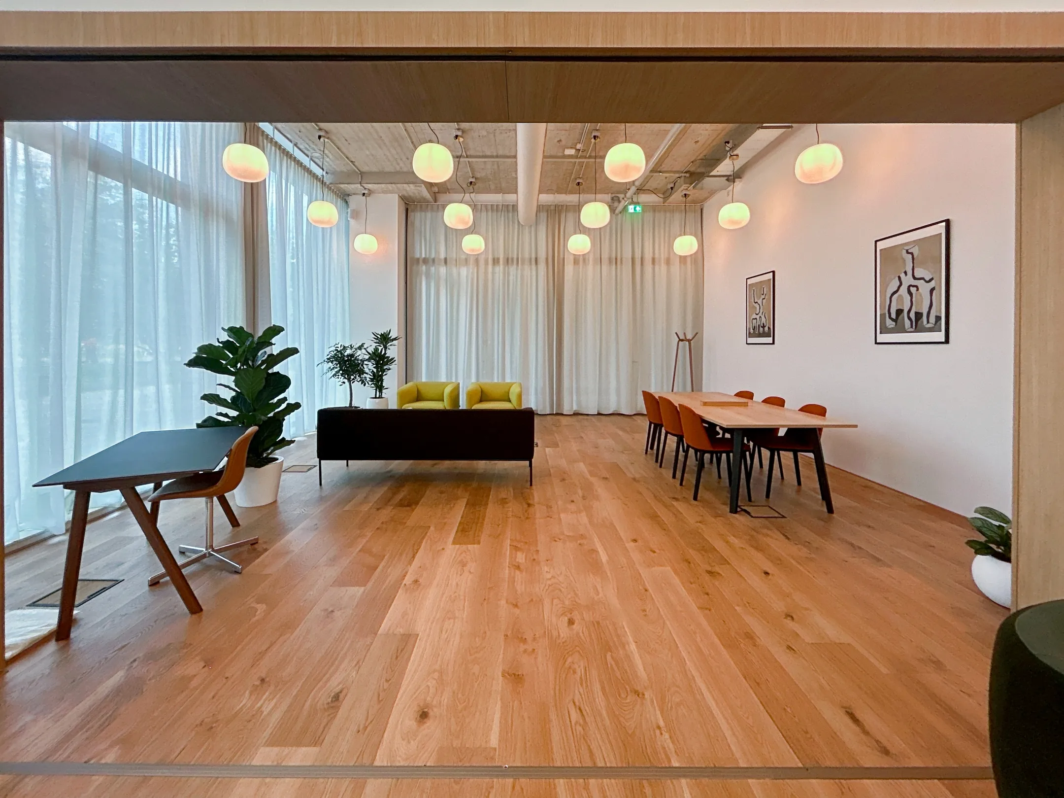 Een moderne vergaderruimte met houten vloer, kleurrijk meubilair, planten en hangende ronde lampen.