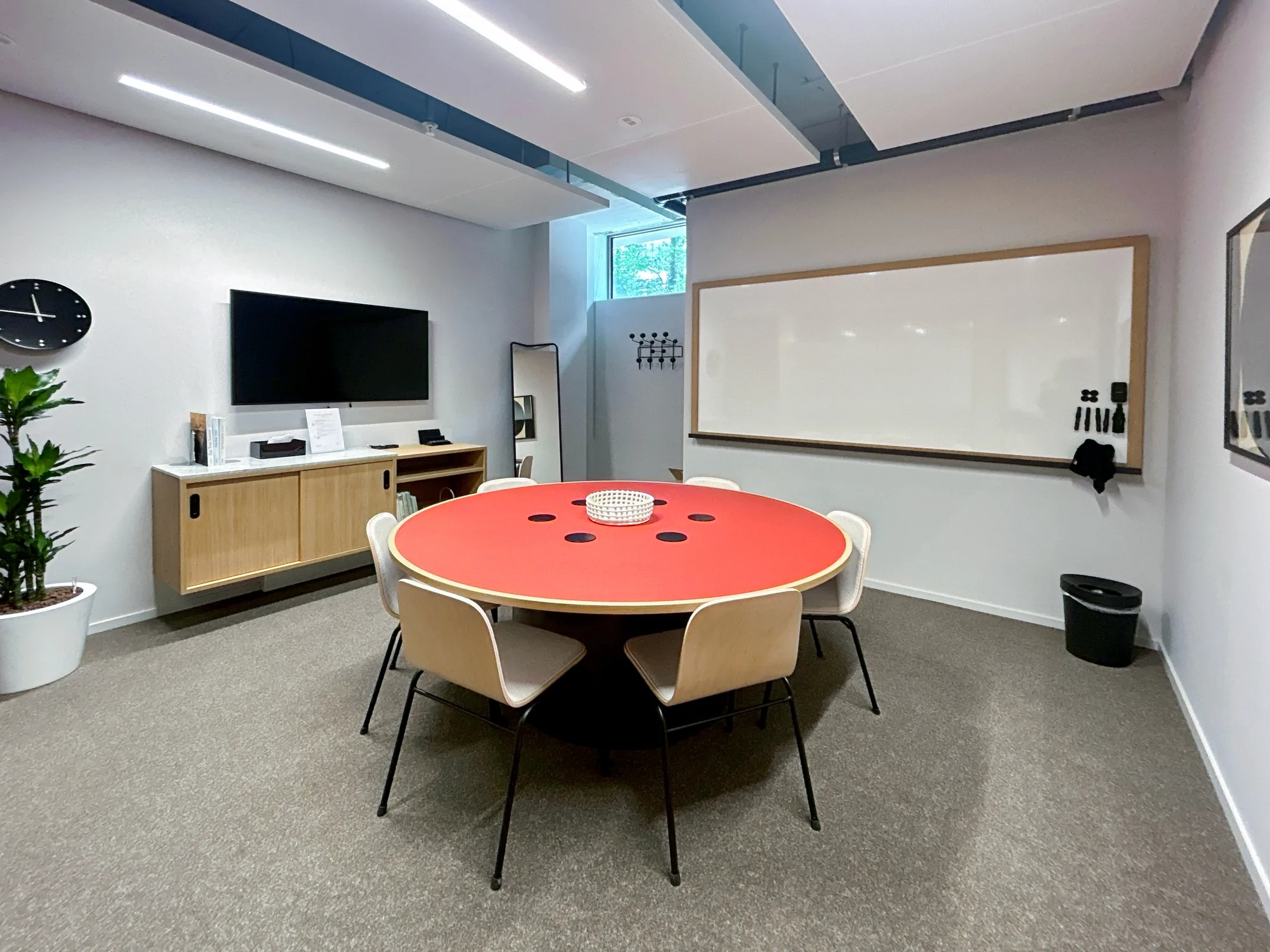 Een moderne vergaderruimte met een ronde rode tafel, stoelen, een whiteboard en een groot scherm aan de muur.