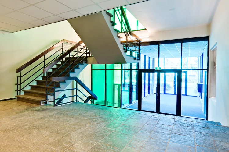 Ingangshal van een modern gebouw aan de Vlaardingenlaan met trap en grote ramen met gekleurde glaspartijen.