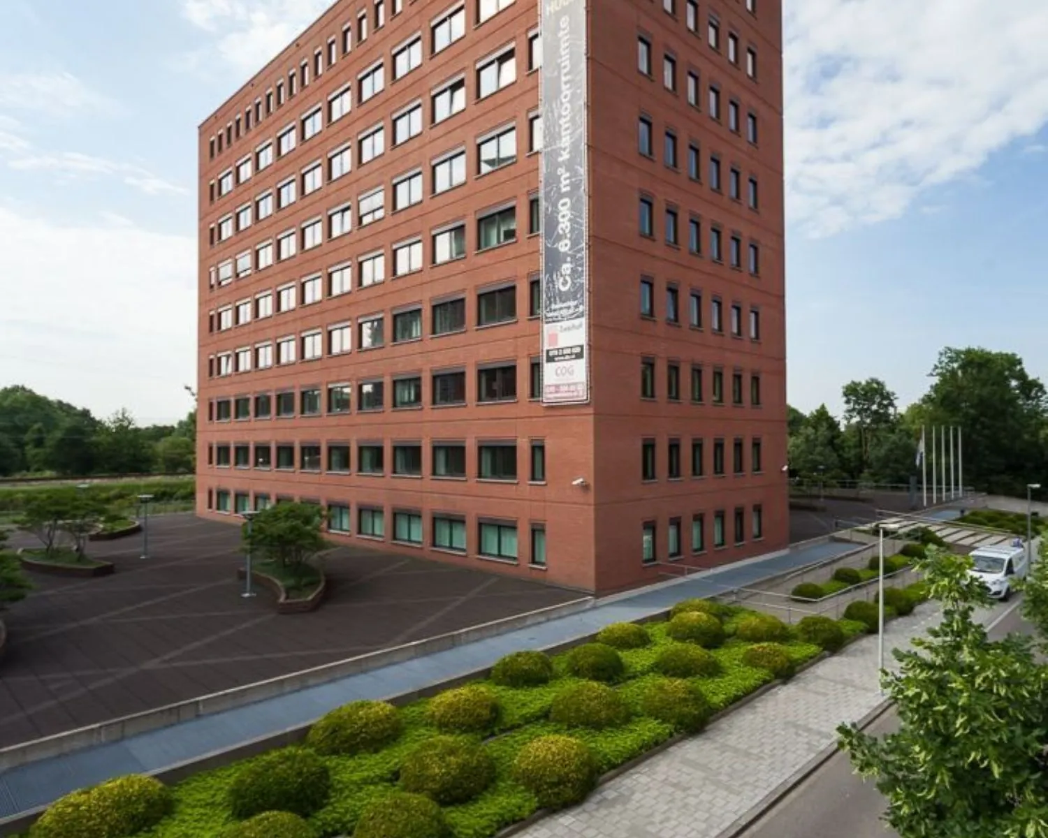 Modern rood bakstenen kantoorgebouw met negen verdiepingen en een groene, verzorgd aangelegde voortuin.