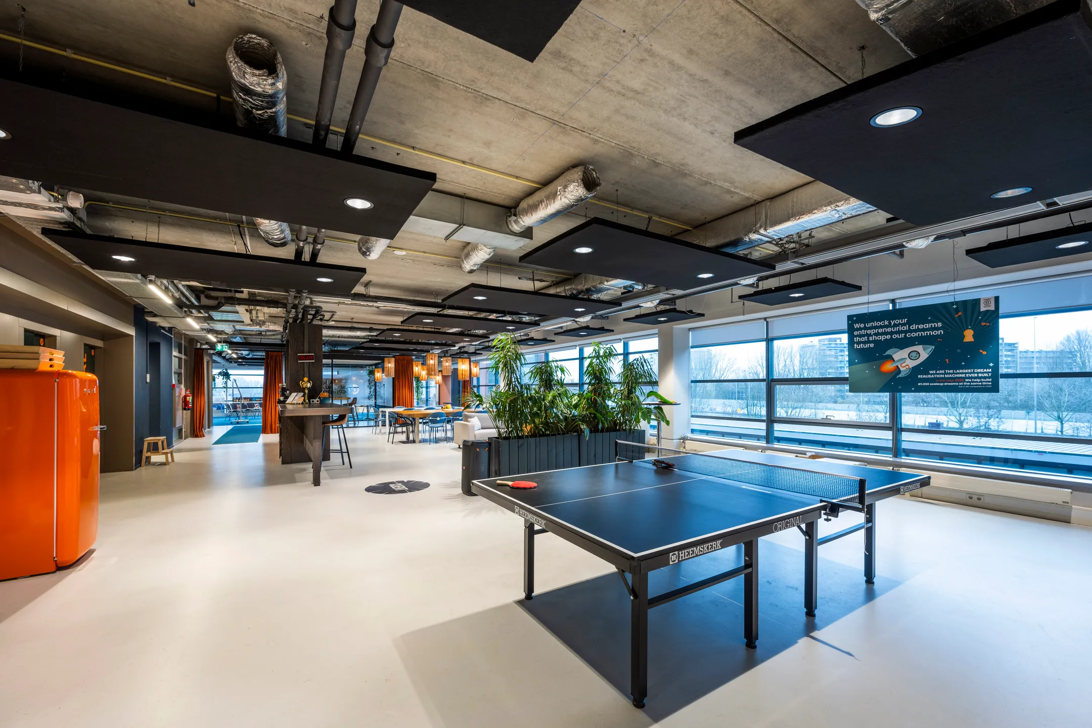 Een open kantoorruimte met een tafeltennistafel, oranje koelkast, en veel natuurlijke lichtinval.