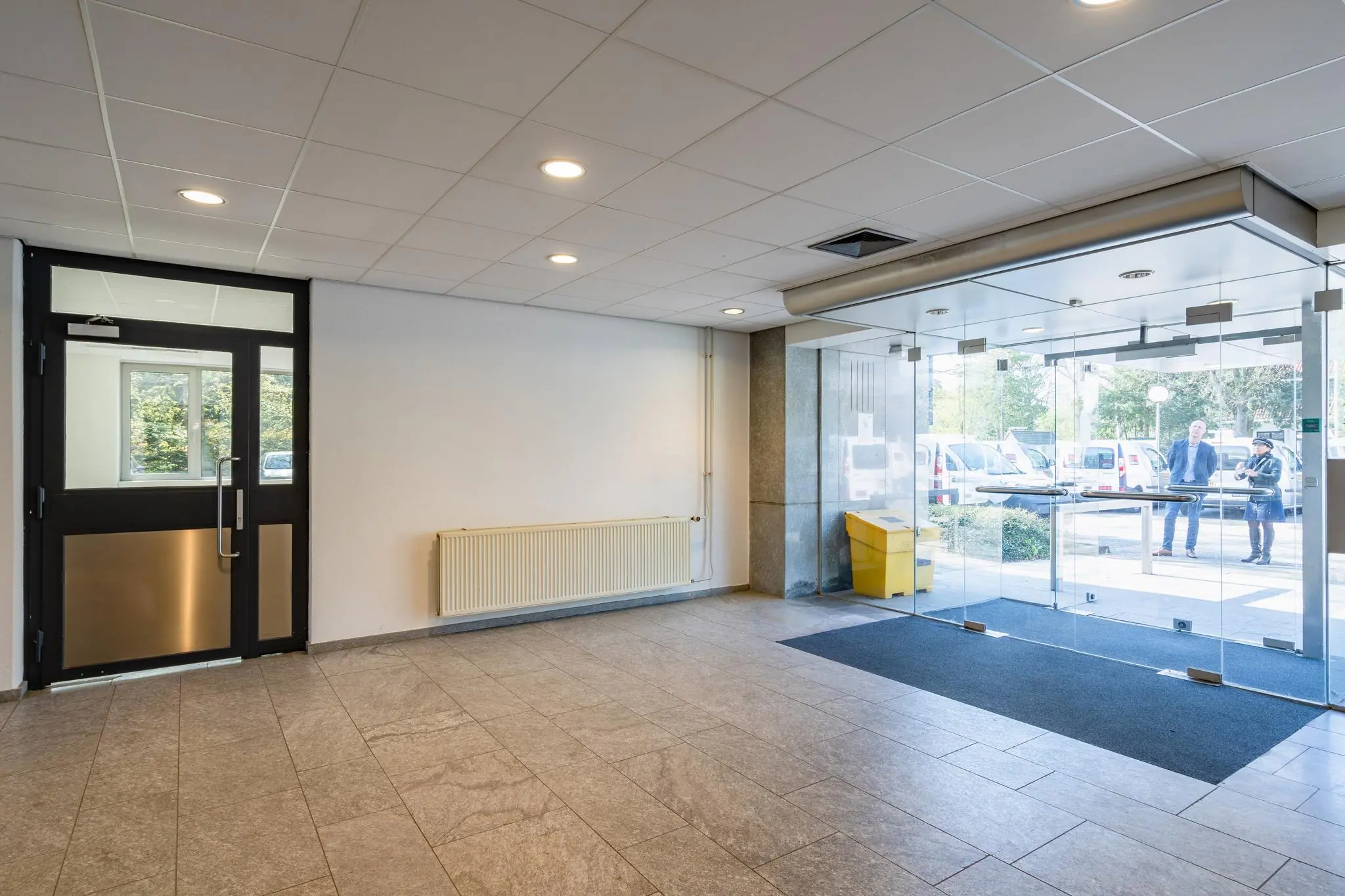 Entreehal met glazen schuifdeuren aan de Utrechtseweg, met zicht op geparkeerde voertuigen en twee mensen buiten.