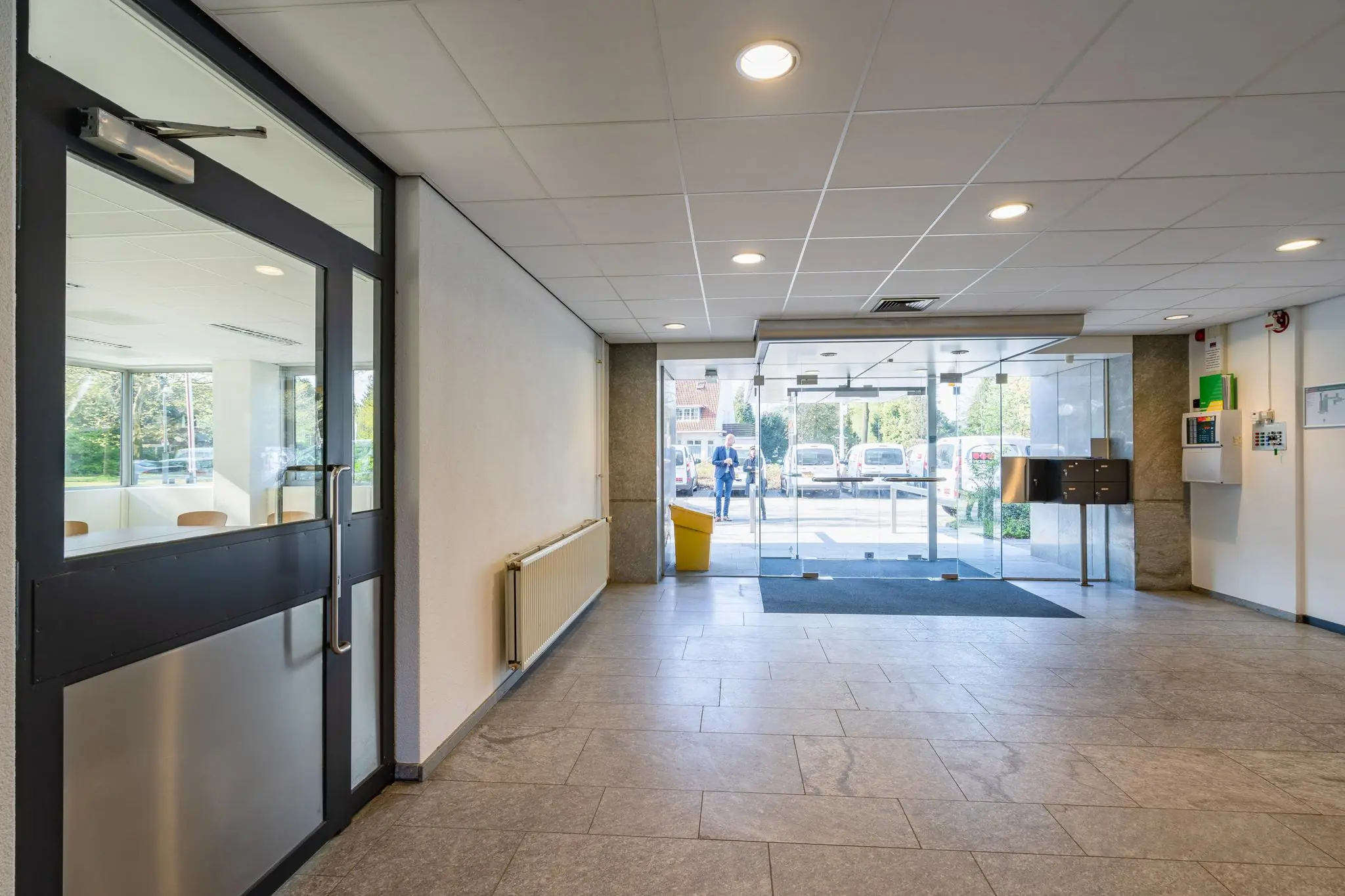 Entreehal van een kantoorpand aan de Utrechtseweg met glazen schuifdeuren en zicht op geparkeerde auto's buiten.