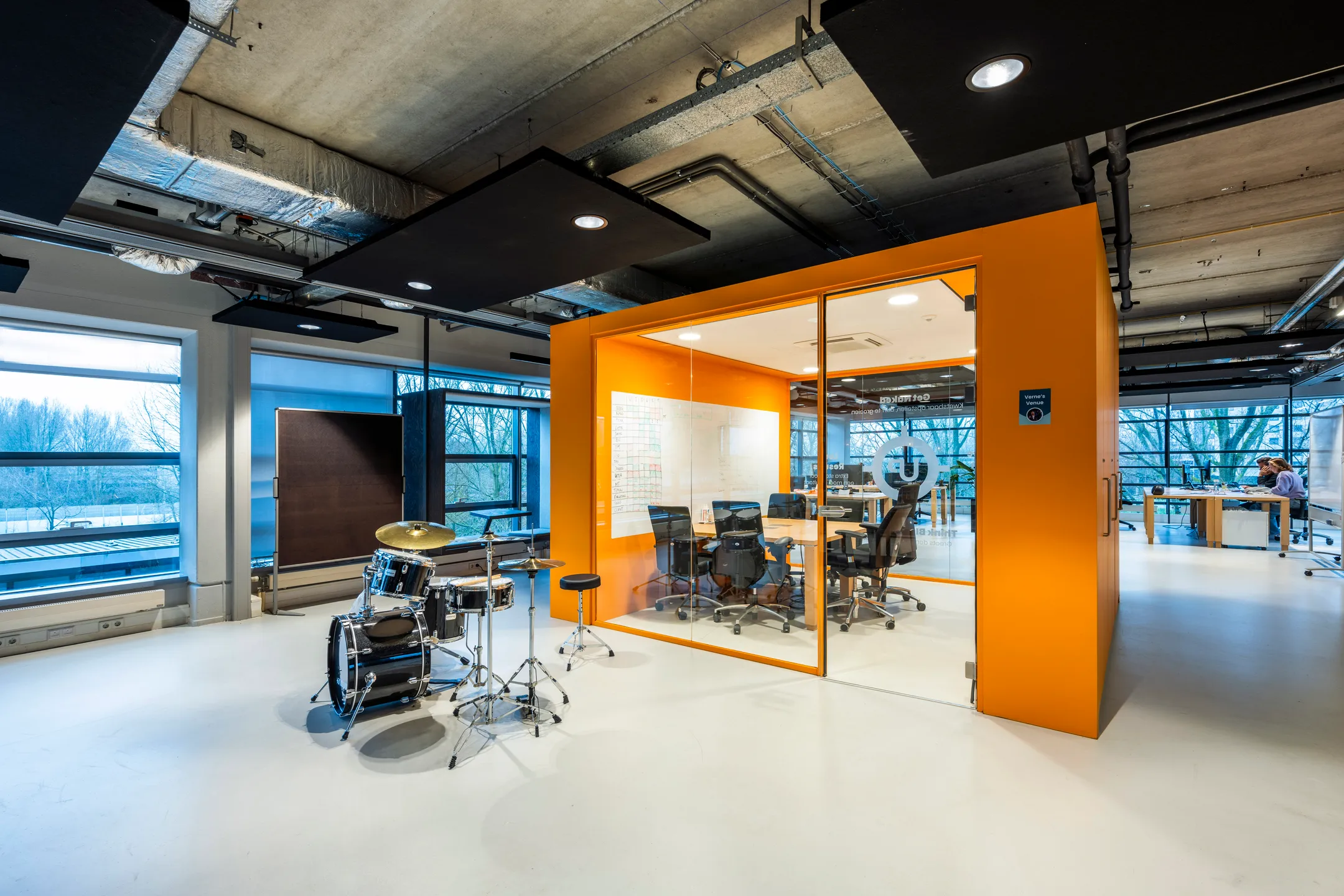 Een moderne kantoorruimte met een oranje glazen vergaderruimte en een drumstel in de hoek.