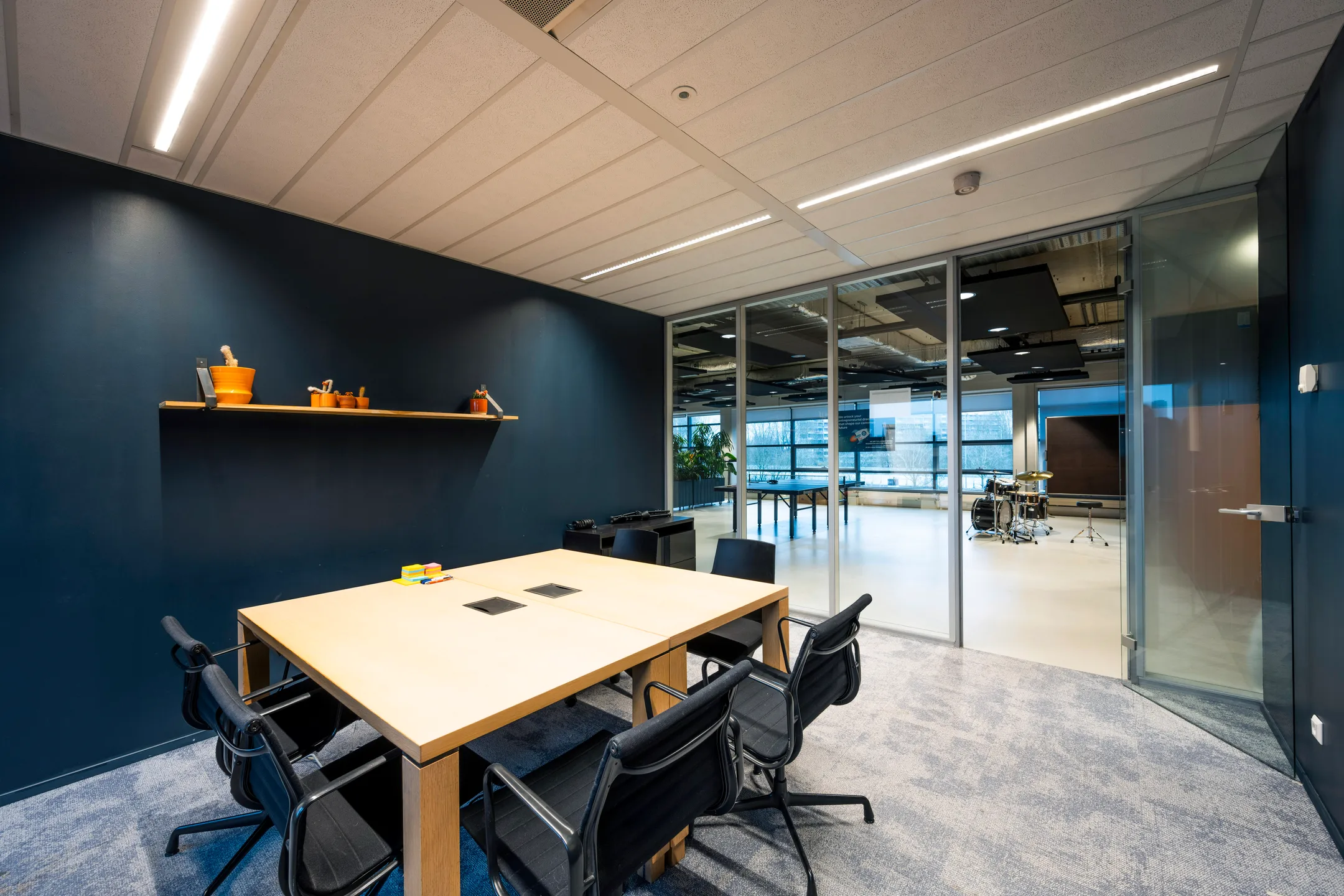 Een moderne vergaderruimte met een houten tafel, zwarte stoelen en glazen wanden die uitkijken op een open ruimte met muziekinstrumenten.