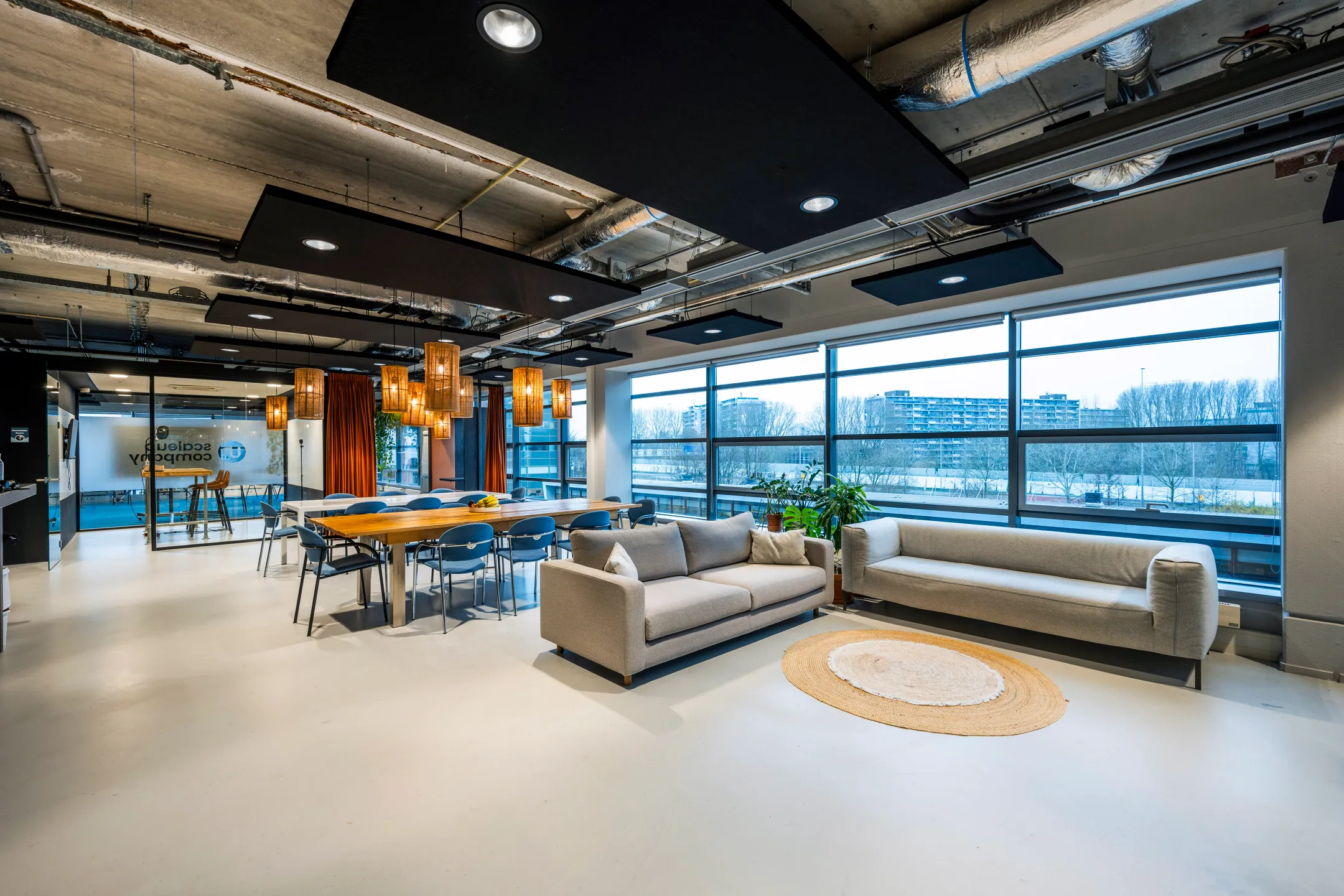 Moderne, open kantoorruimte met zitgedeelte en vergadertafel, omringd door grote ramen en industriële plafonddetails.