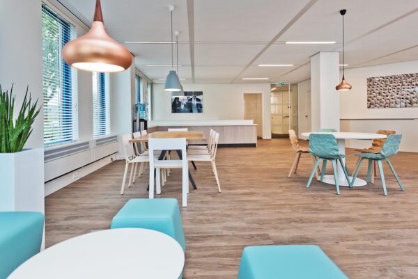 Moderne en lichte kantoorkantine met houten vloer, kleurrijke stoelen en hanglampen aan het plafond.