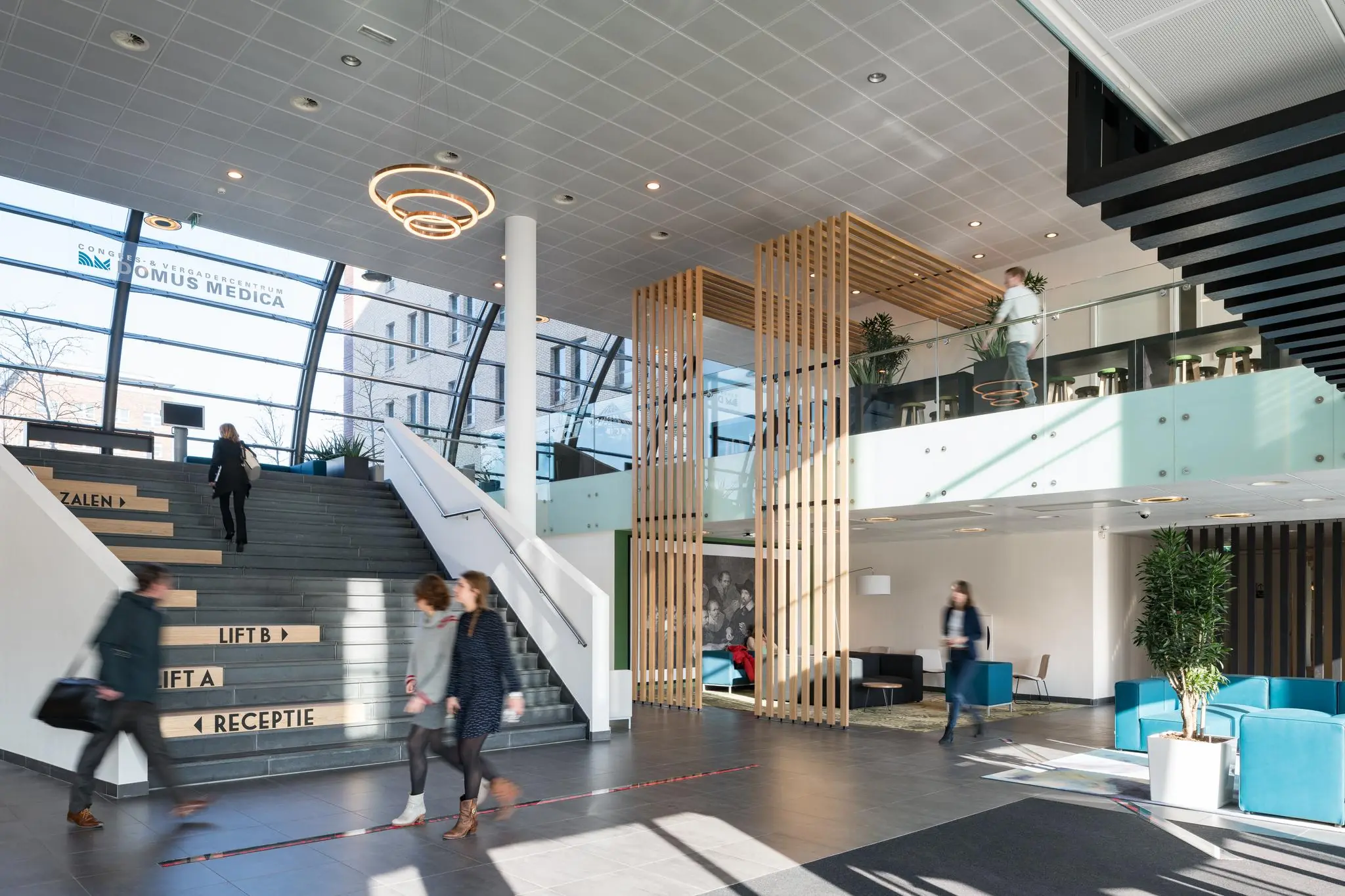 Moderne en lichte entreehal van Domus Medica aan de Mercatorlaan met trap, zitgedeelte en passerende mensen.
