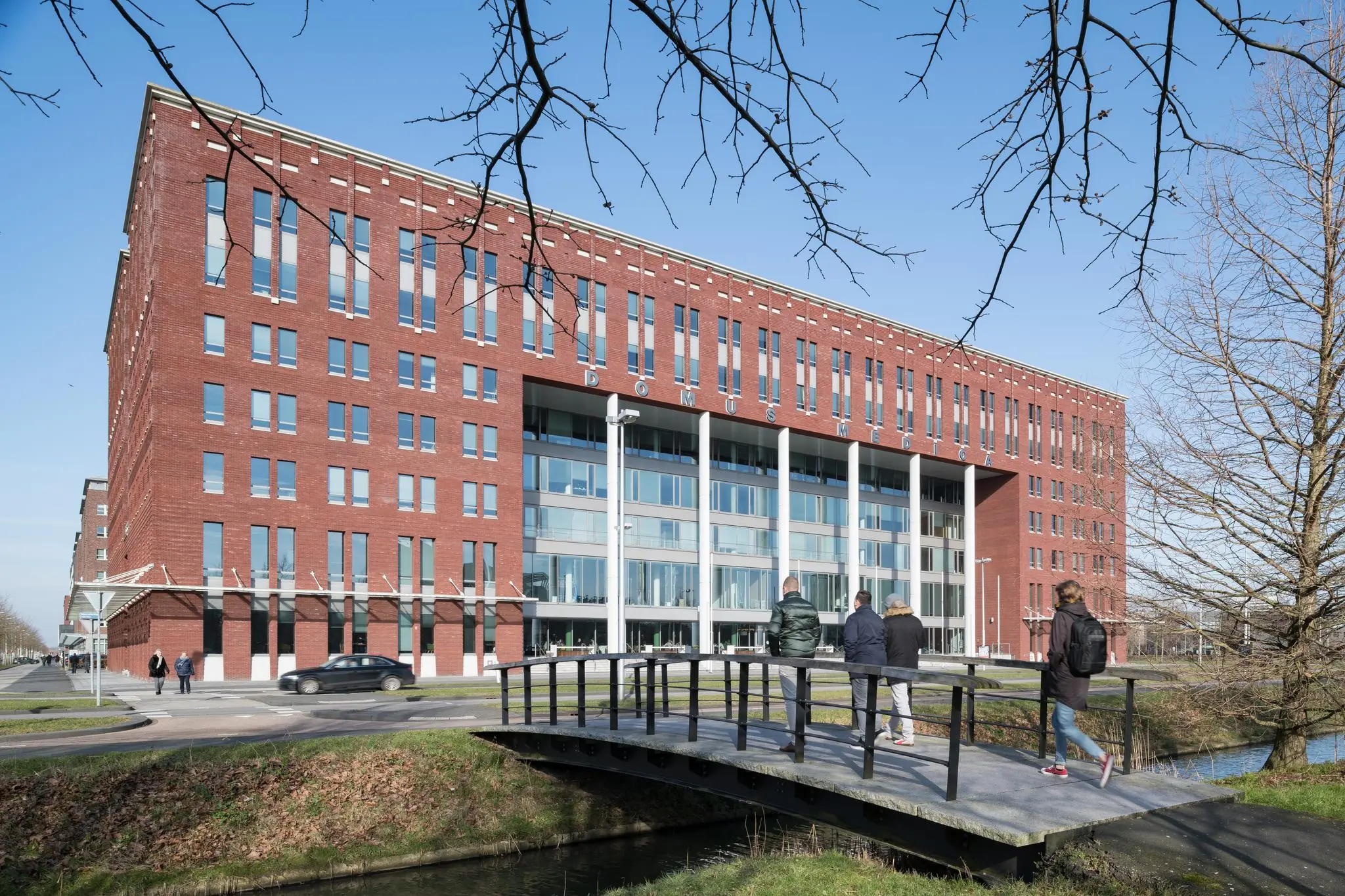 Vooraanzicht van het kantoorgebouw Domus Medica aan de Mercatorlaan in Utrecht, met mensen die over een brug lopen.