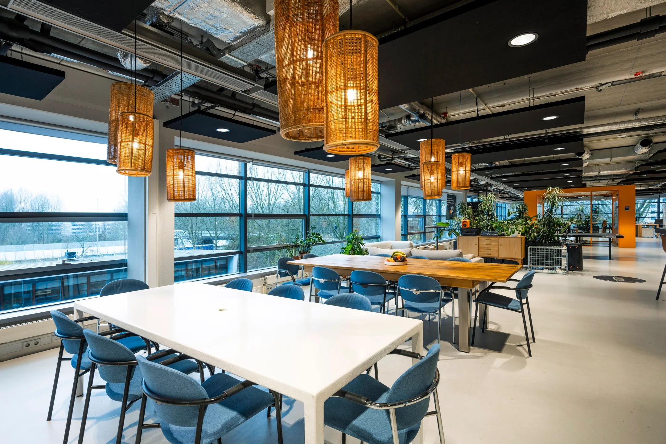 Een moderne kantoorruimte met grote ramen, houten tafels en hangende rieten lampen.