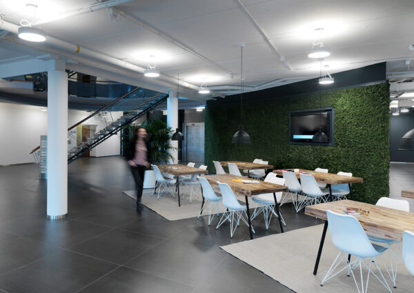 Moderne gemeenschappelijke werkruimte met houten tafels, witte stoelen, een groene moswand en een trap op de achtergrond.