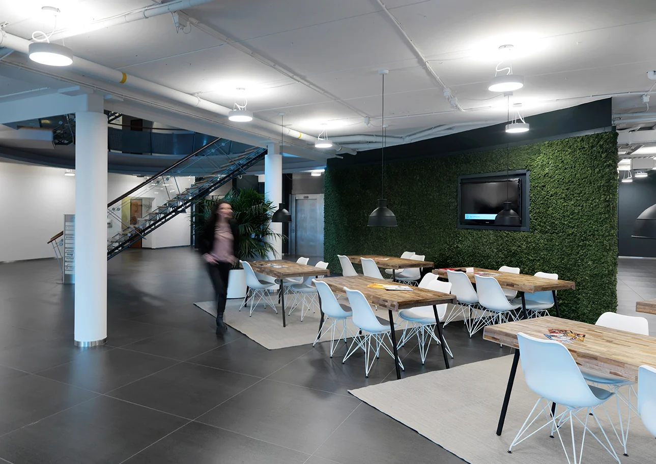 Moderne gemeenschappelijke werkruimte met houten tafels, witte stoelen, een groene moswand en een trap op de achtergrond.