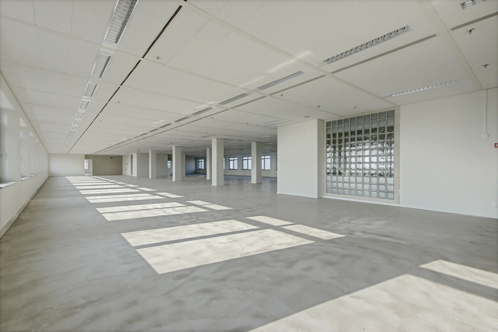 Lichte, lege kantoorruimte aan de Winthontlaan met betonnen vloer, glazen bouwstenen en grote ramen.