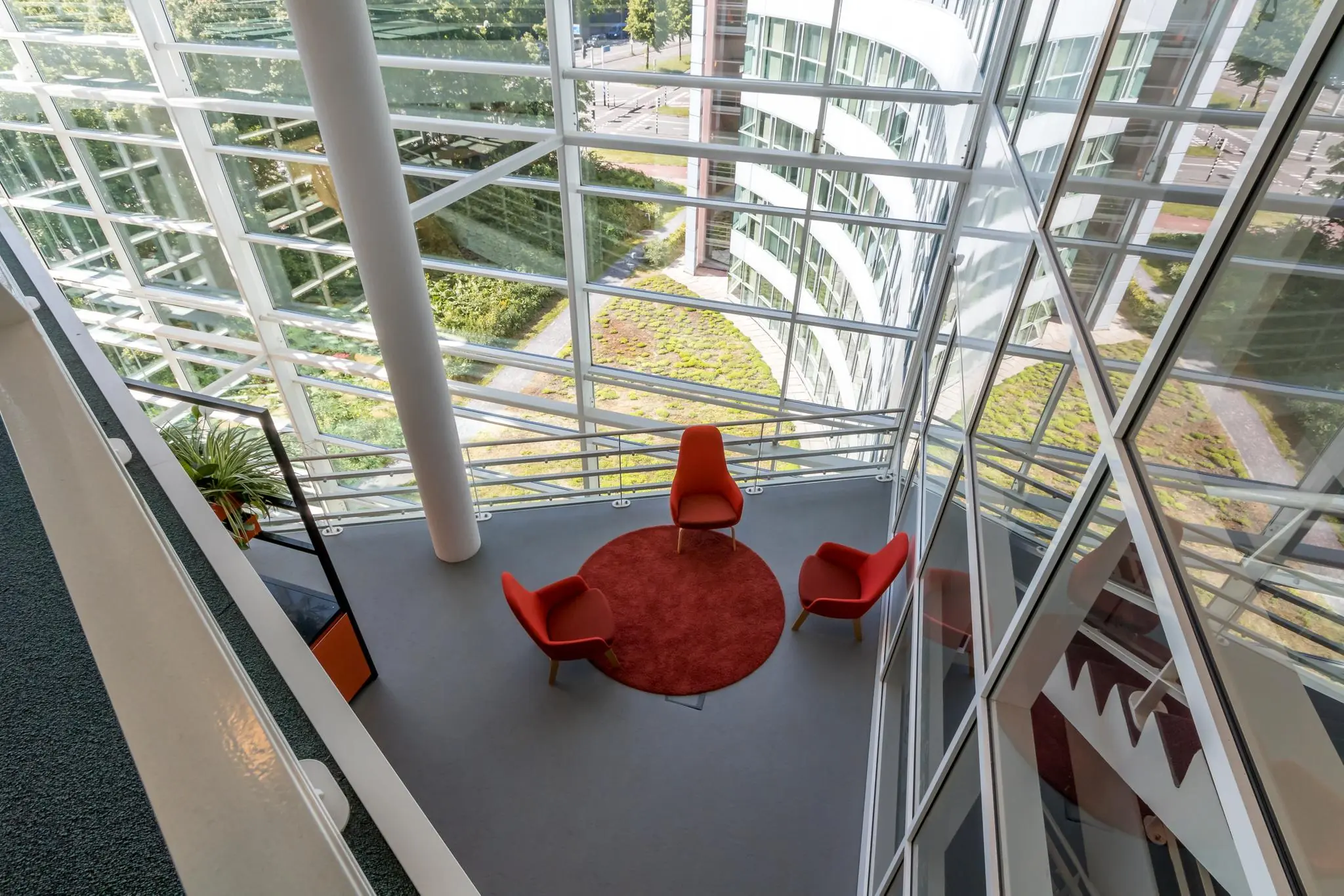 Zithoek met drie rode stoelen en een rood vloerkleed in een glazen atrium van een modern kantoorgebouw aan de Newtonlaan.