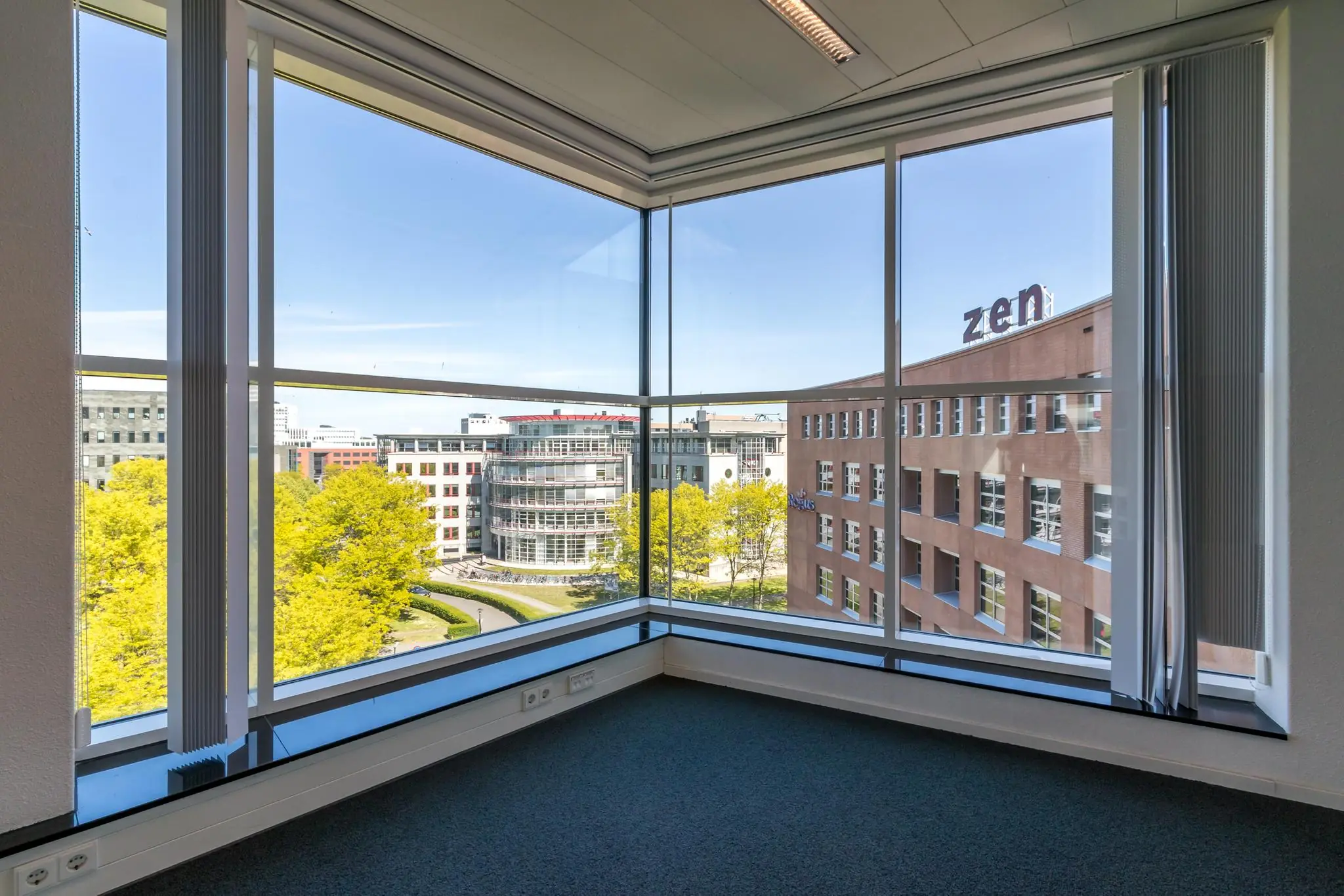 Hoek van een leeg kantoor met grote ramen en uitzicht op kantoorgebouwen aan de Newtonlaan.