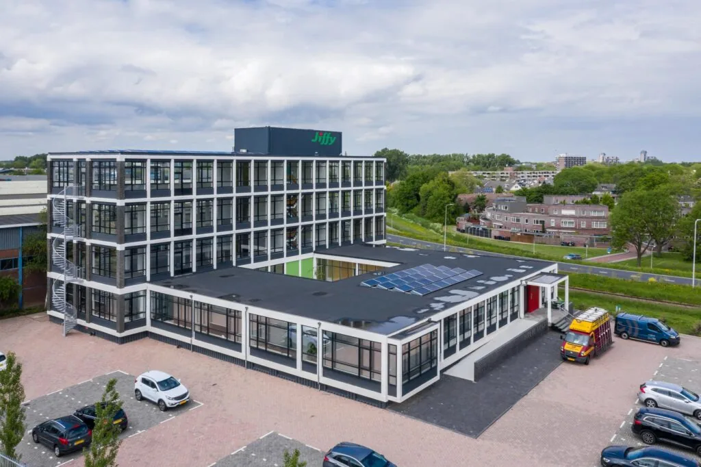 Luchtfoto van het Jiffy-kantoorgebouw aan de Lindtsedijk met zonnepanelen op het dak en geparkeerde voertuigen op de voorgrond.