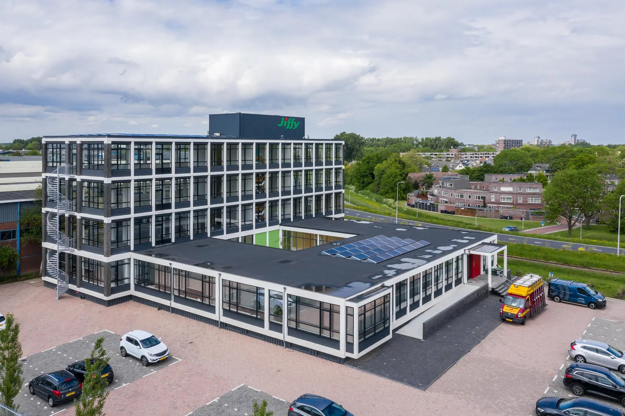 Luchtfoto van het Jiffy-kantoorgebouw aan de Lindtsedijk met zonnepanelen op het dak en geparkeerde voertuigen op de voorgrond.