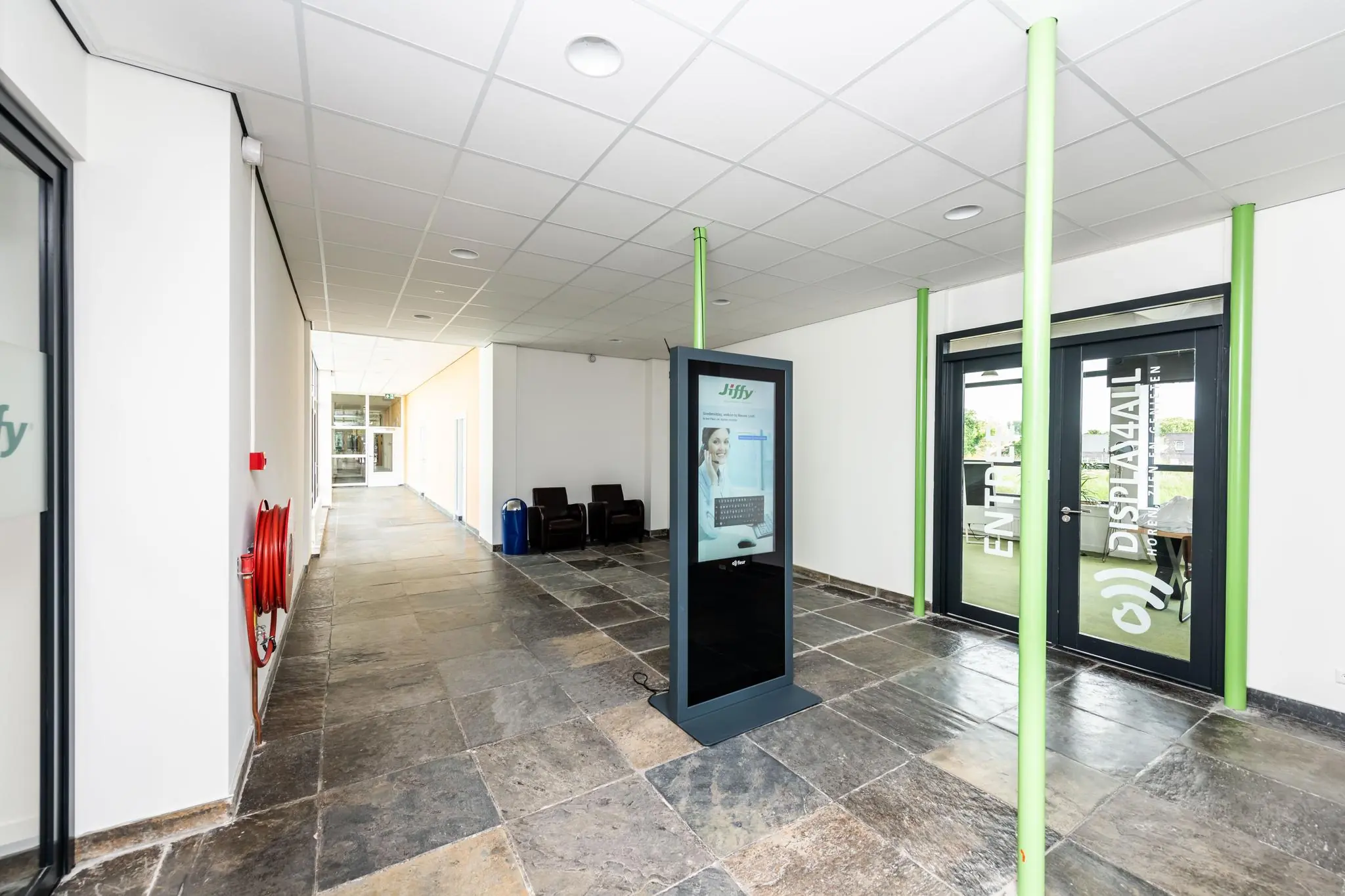 Ingang van het kantoorpand aan de Lindtsedijk met een digitale zuil, glazen deuren van Display4All en een brede gang met stoelen.