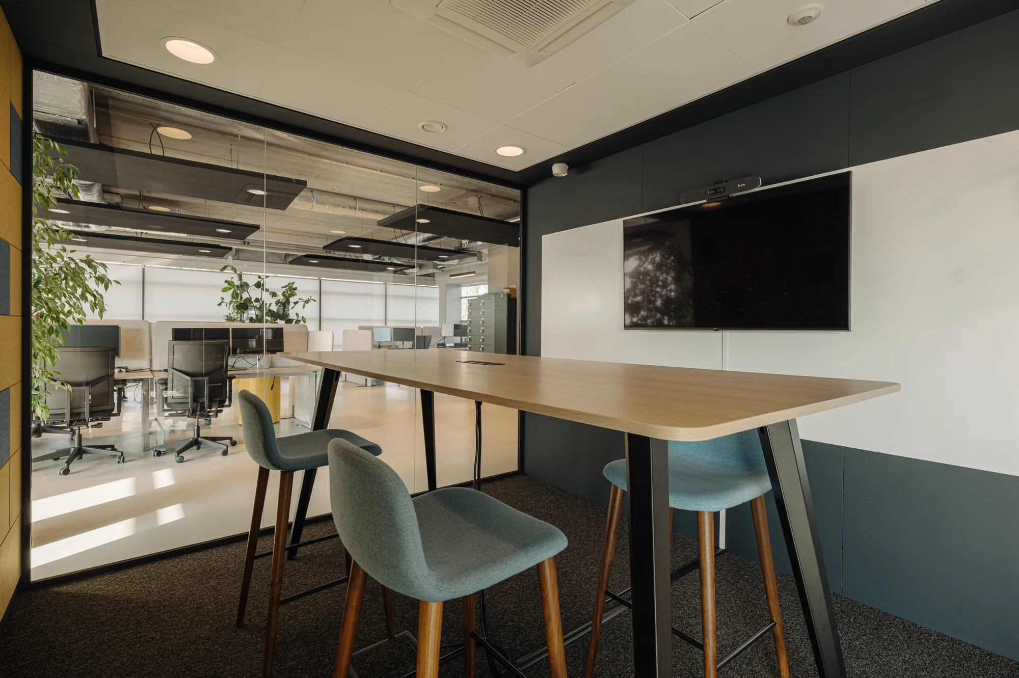 Moderne kantoorruimte met een hoge tafel en stoelen, naast een glazen wand met zicht op bureaus en bureaustoelen.
