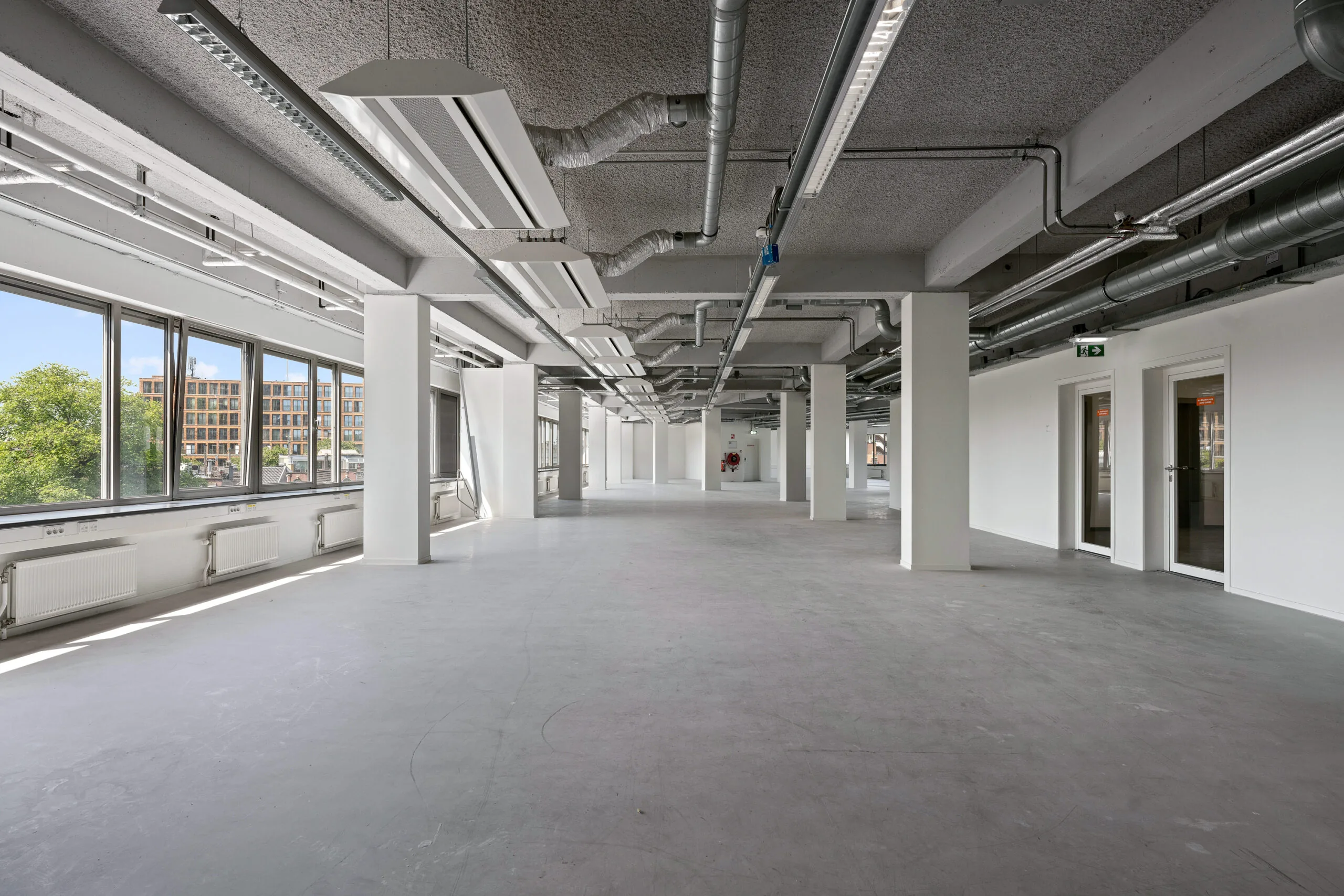 Lichte, lege kantoorruimte met industriële afwerking en grote ramen aan de linkerkant.