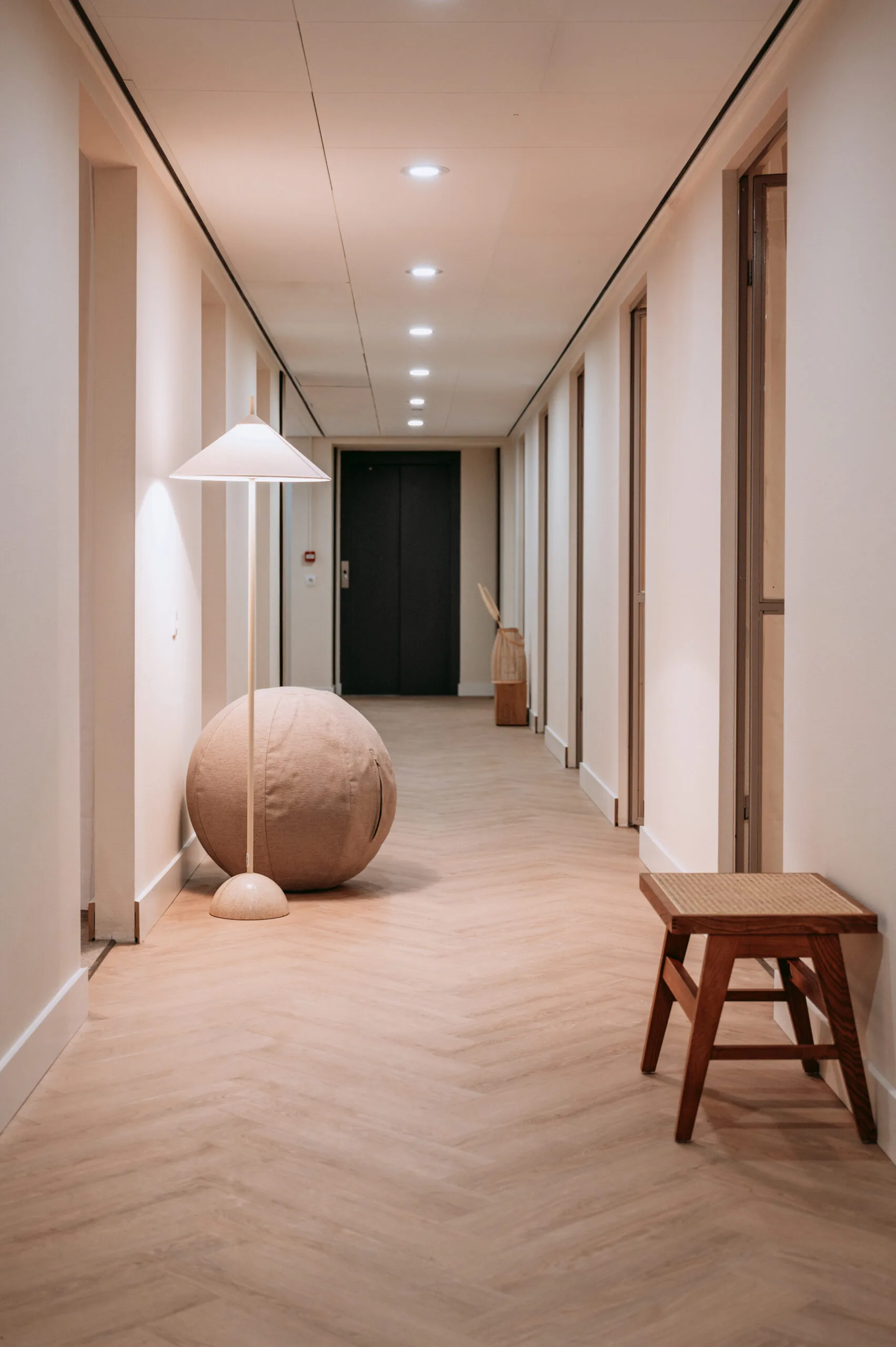 Stijlvol verlichte gang met houten vloer, designlamp, ronde poef en houten krukje.