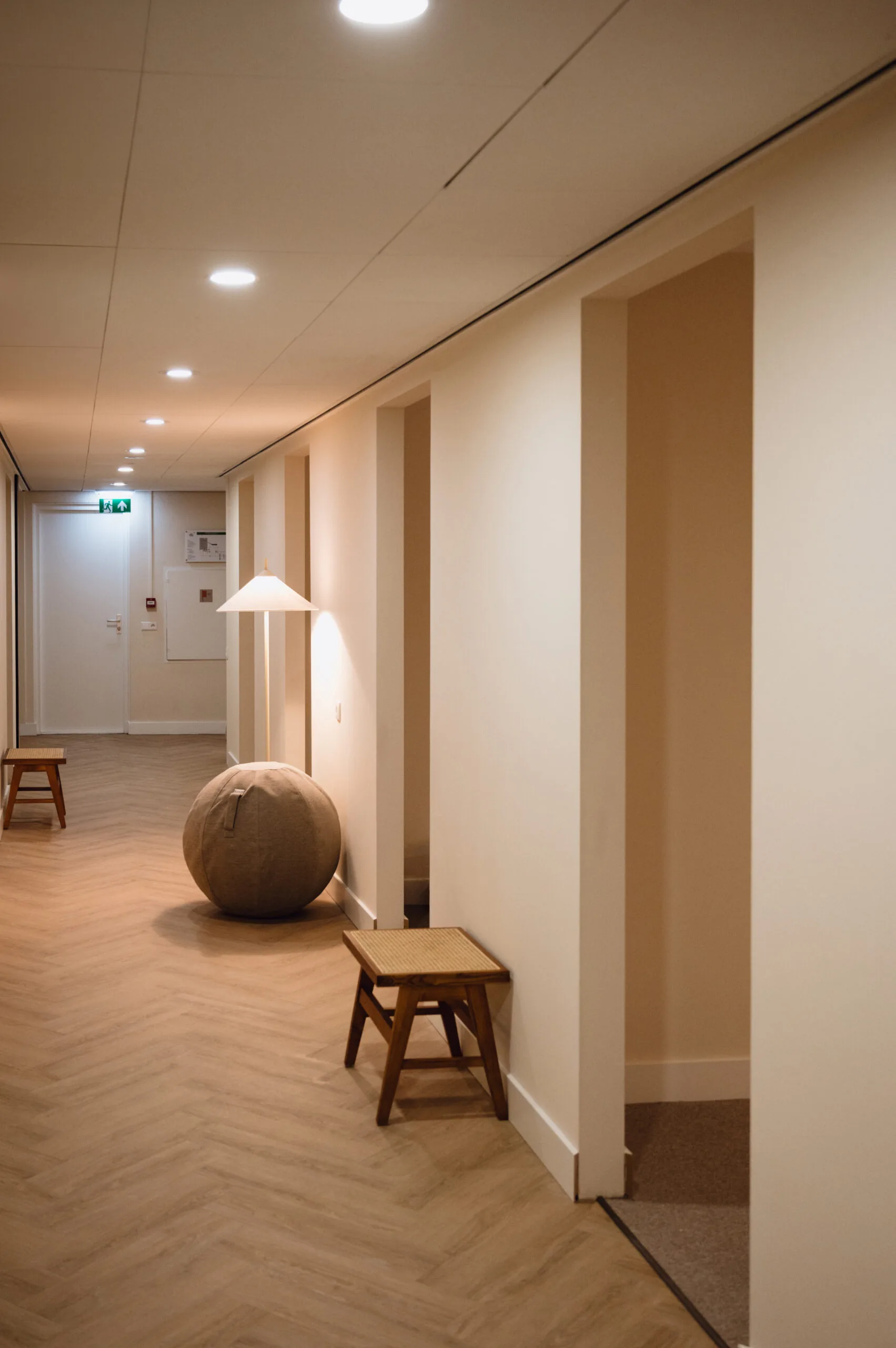 Verlichtte gang met houten vloer, krukjes en een ronde lamp naast een deur.