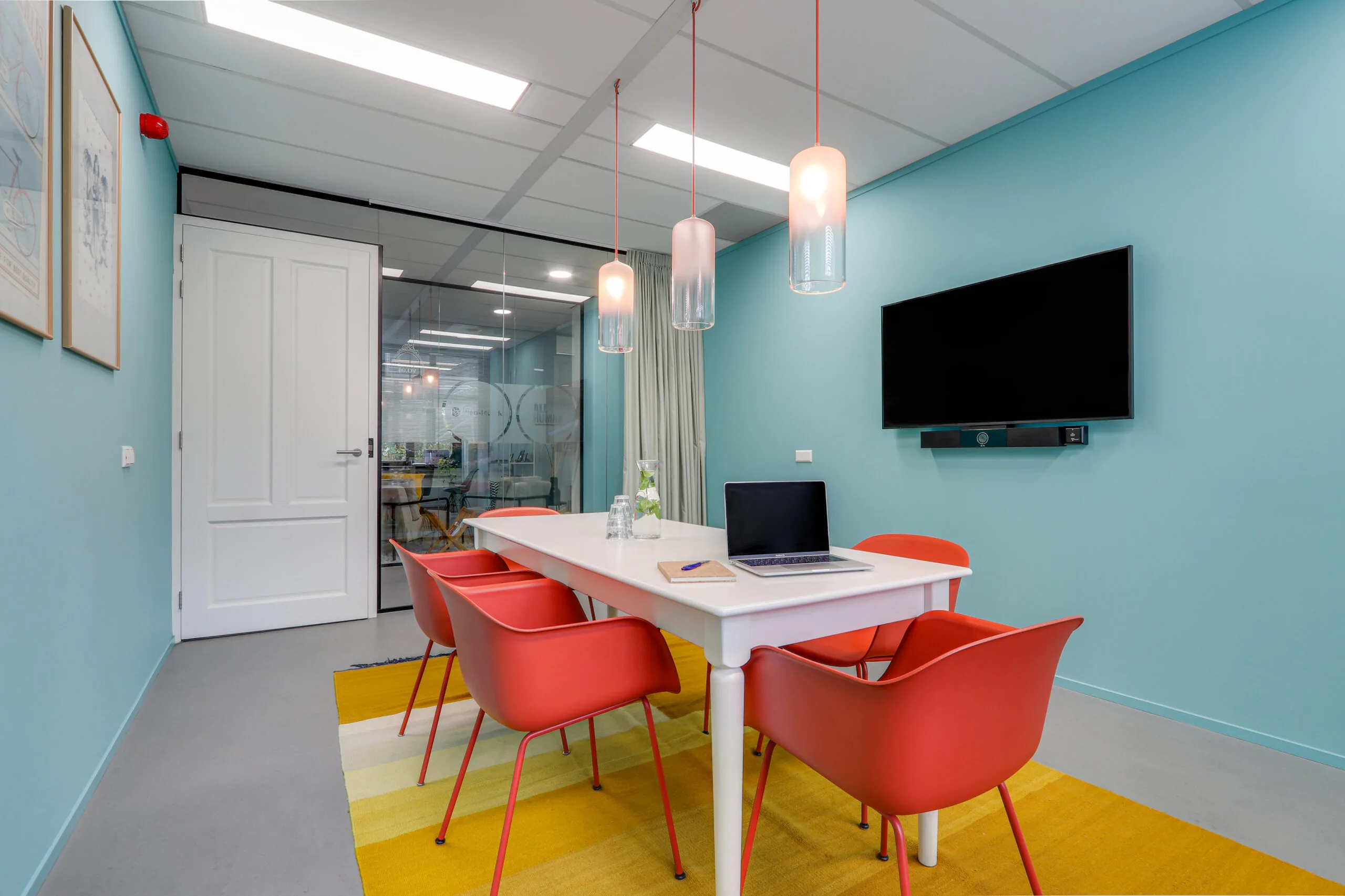 Vergaderruimte in Rotterdam Centrum met rood-oranje stoelen, witte tafel, wandtelevisie en glazen wand.