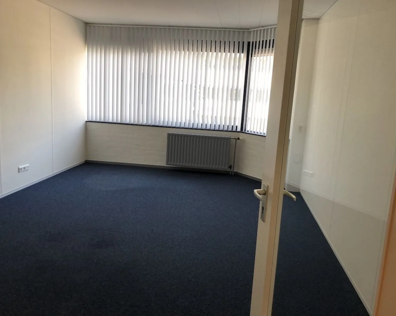 Lege kantoorruimte met blauw tapijt en grote ramen met verticale lamellen aan de Hooilaan 1 in Breda.