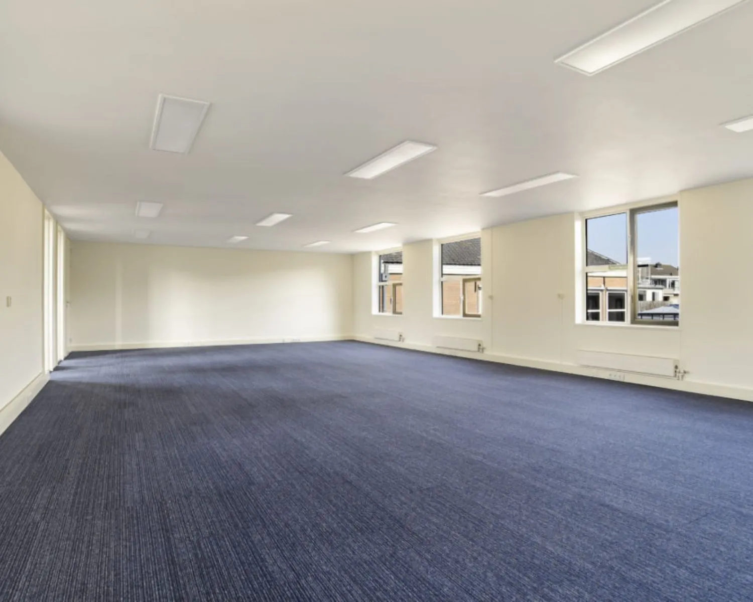 Lichte, ruime kantoorruimte met blauw tapijt en grote ramen aan de Wilhelminastraat 43A in Haarlem.