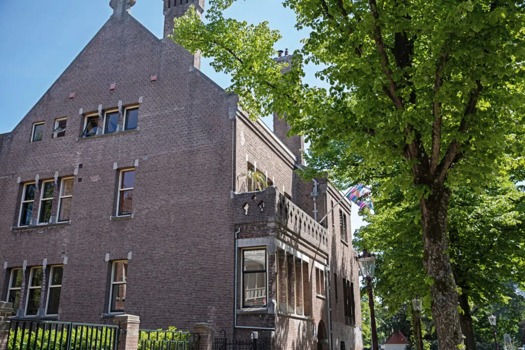 Bakstenen gebouw met meerdere ramen en een boom aan een zonnige straatkant.