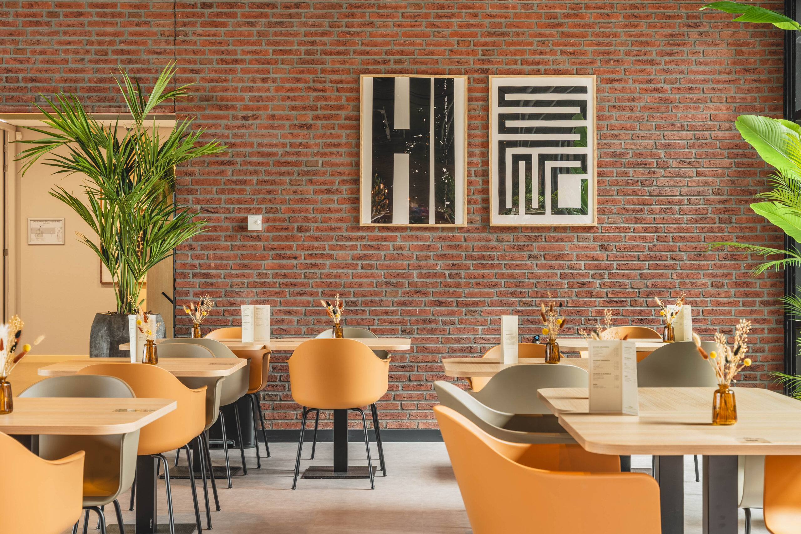 Een modern ingerichte eetruimte met oranjerode stoelen, houten tafels, groene planten en twee kunstwerken aan een bakstenen muur.