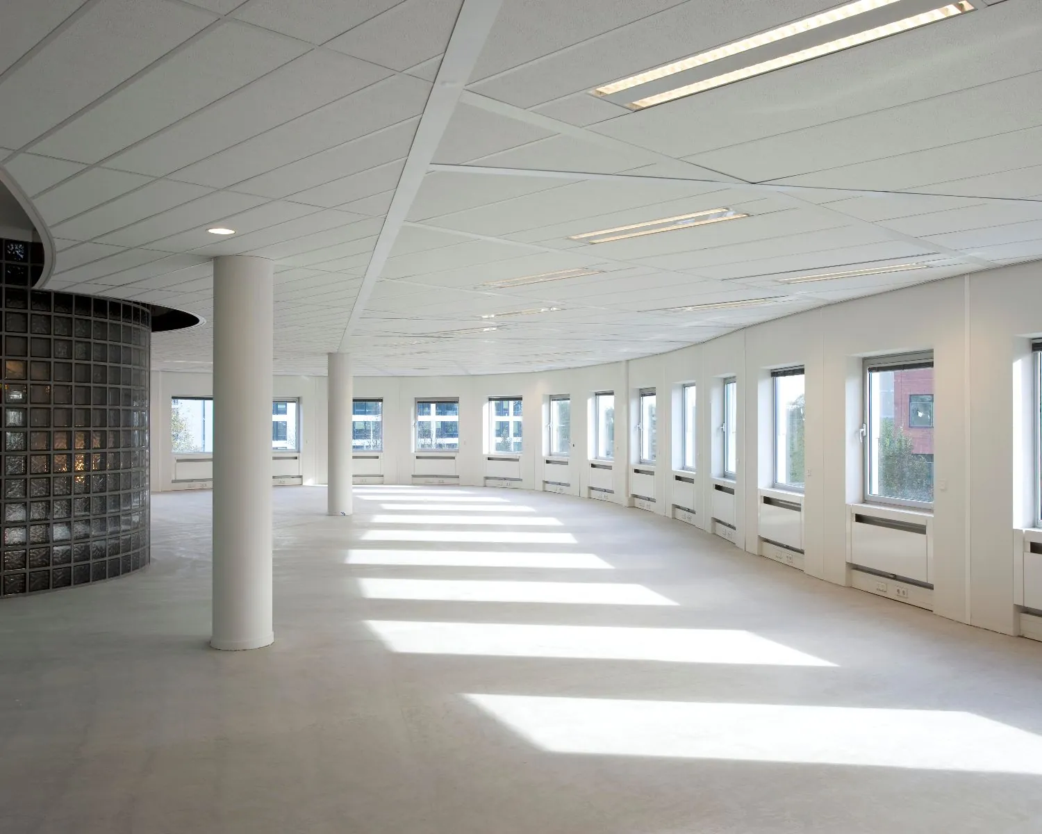 Lichte, lege kantoorruimte met gebogen glazen wand en rijen ramen langs de buitenmuur.