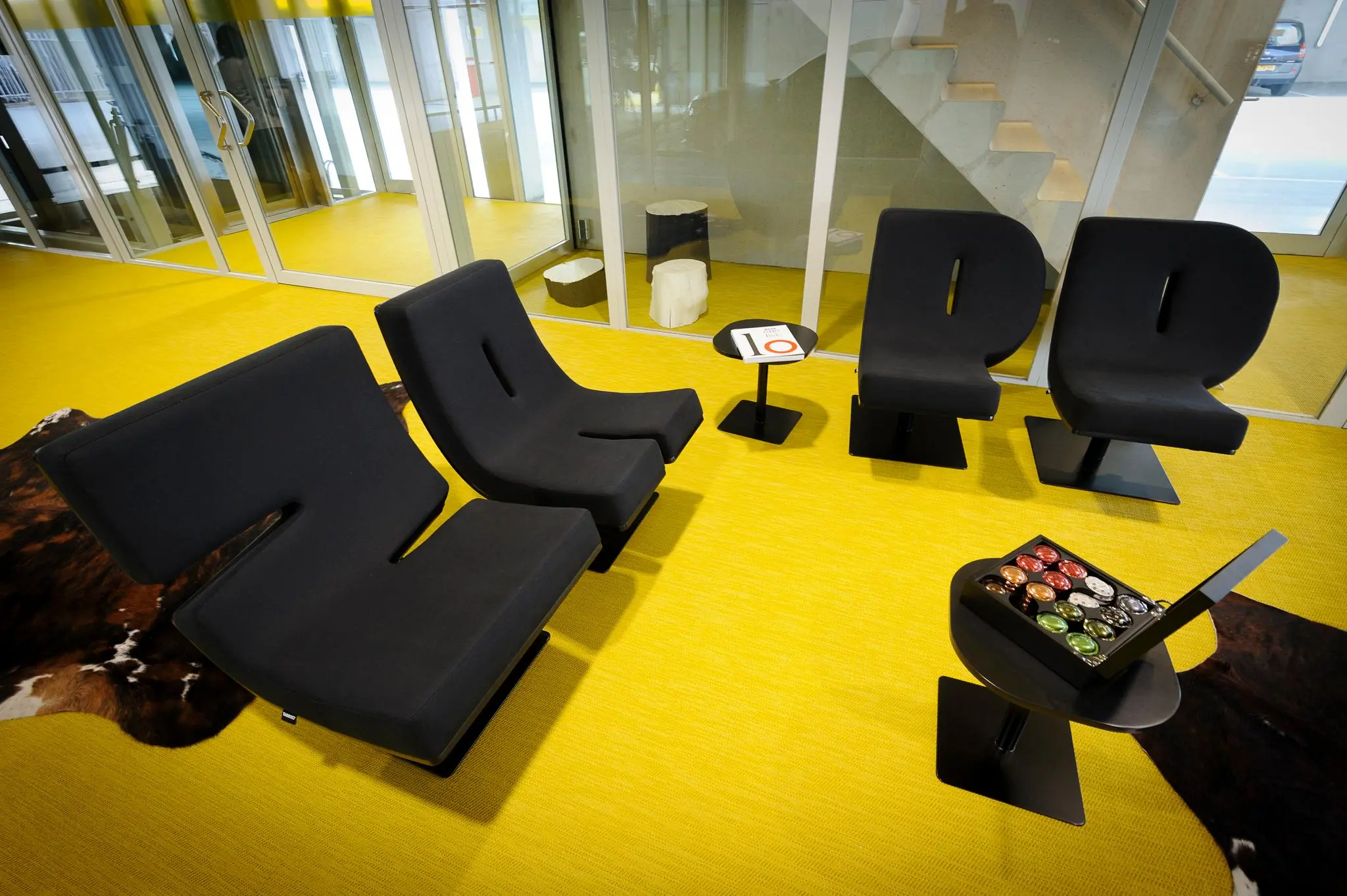 Moderne wachtruimte aan de Bahialaan met zwarte designstoelen, felgele vloerbedekking en een tafeltje met koffiecups.