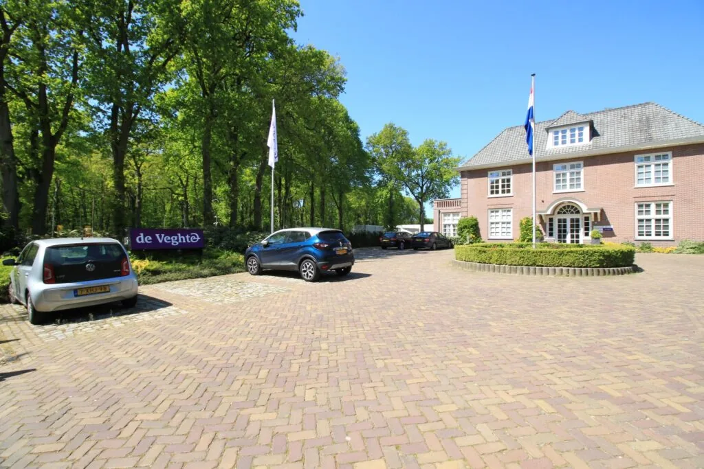 Vooraanzicht van een statig kantoorpand aan de Amersfoortsestraat met oprijlaan, geparkeerde auto's en een bord met de naam 'de Veghte'.