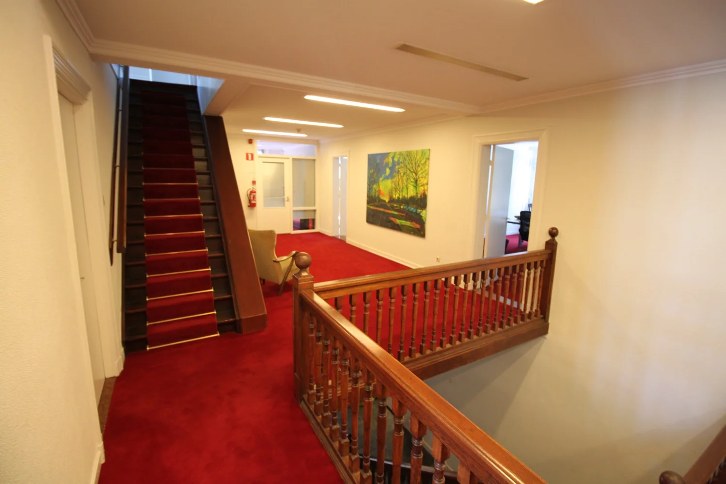 Binnenhal met rode vloerbedekking, houten trap en balustrade, kleurrijk schilderij aan de muur en meerdere deuren naar aangrenzende kamers.
