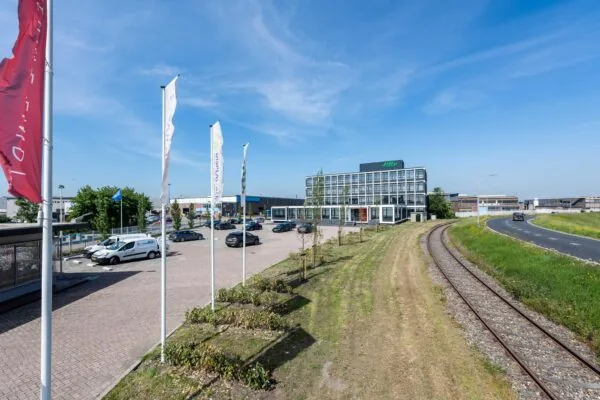 Kantoor van Jiffy aan de Lindtsedijk met parkeerplaats, vlaggenmasten en een spoorlijn langs de weg op een zonnige dag.