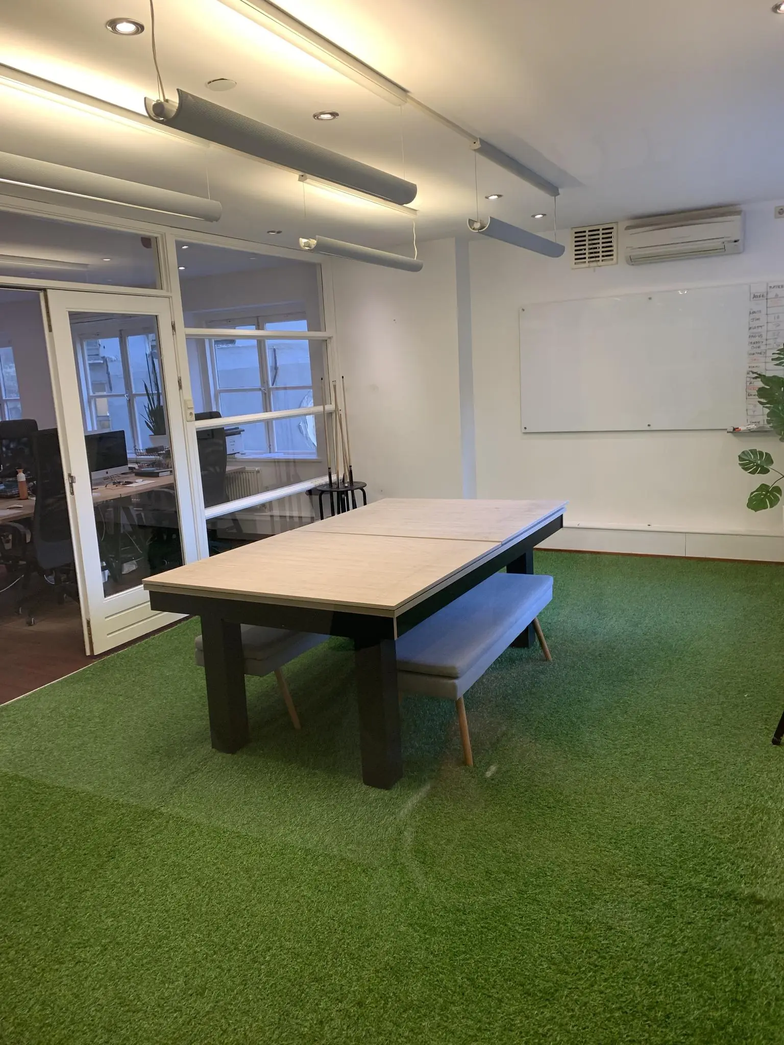 Vergaderruimte aan de Keizersgracht met kunstgrasvloer, houten tafel, banken en glazen wand naar naastgelegen kantoor.