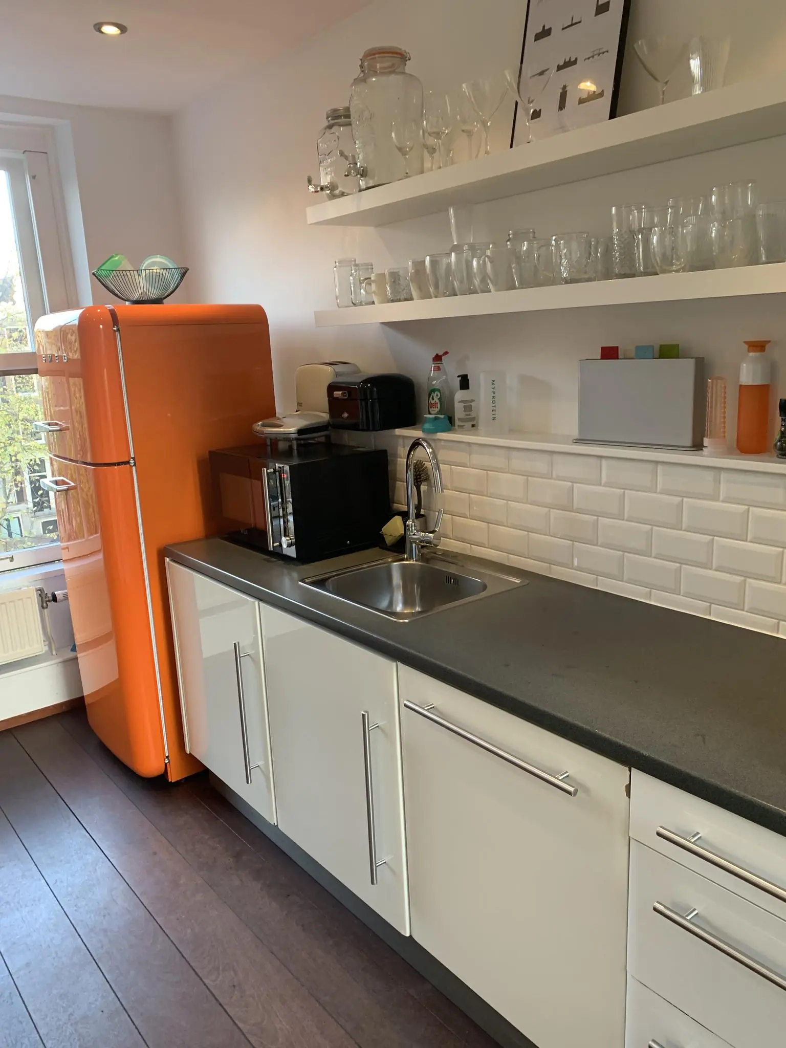 Moderne keuken met een oranje retro koelkast, witte kasten en open planken vol glazen.