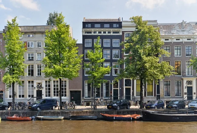 Gevels van historische grachtenpanden aan de Keizersgracht in Amsterdam met bomen, auto's en boten langs het water.
