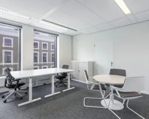 Moderne, lichte kantoorruimte met twee bureaus, een ronde vergadertafel en uitzicht op een klassiek gebouw.