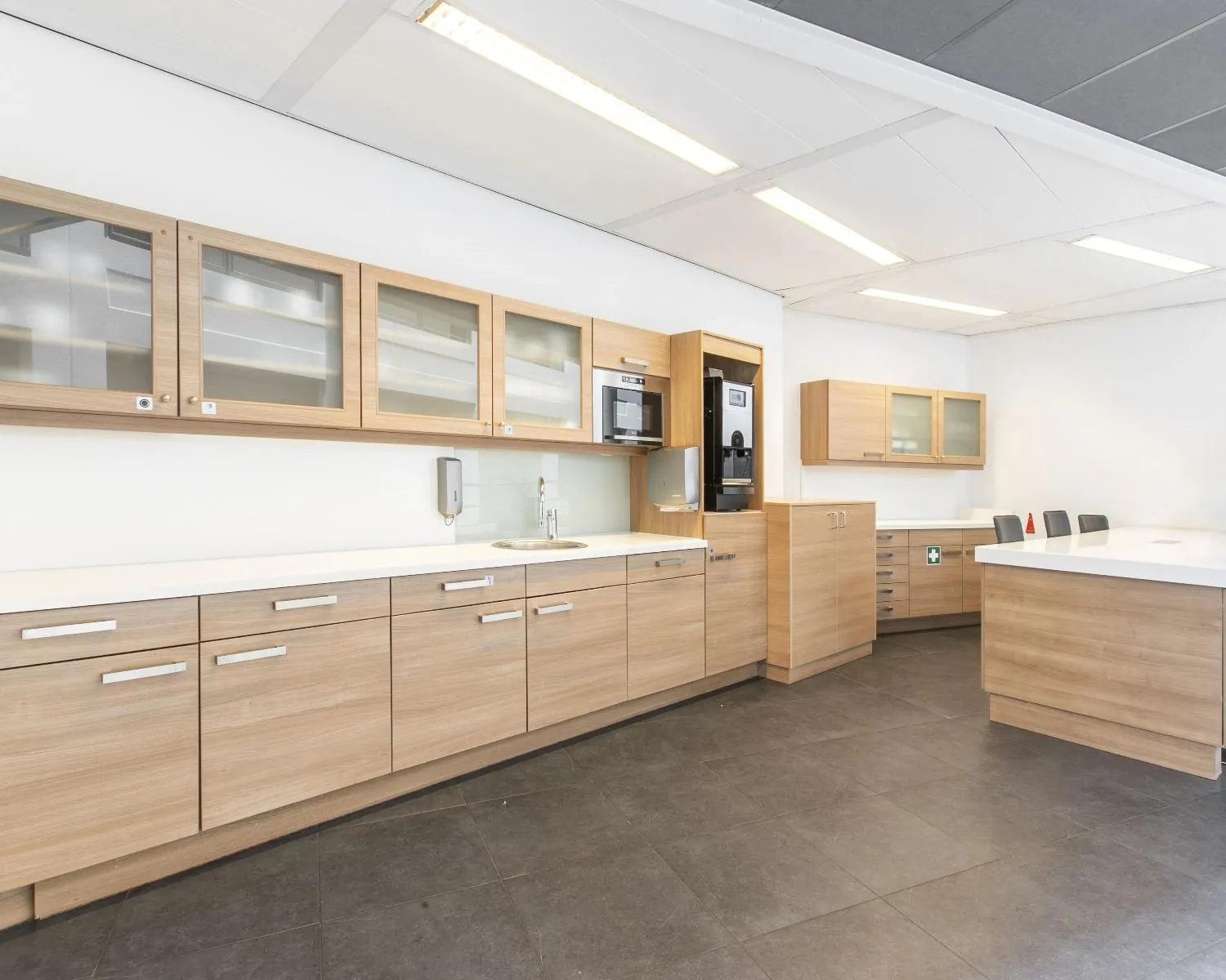 Moderne pantrykeuken met houtkleurige kasten, witte werkbladen en ingebouwde koffiemachine in een kantooromgeving.