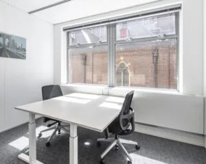 Licht en modern kantoor met twee bureaustoelen, een wit bureau en uitzicht op een oud bakstenen gebouw met glas-in-loodraam.