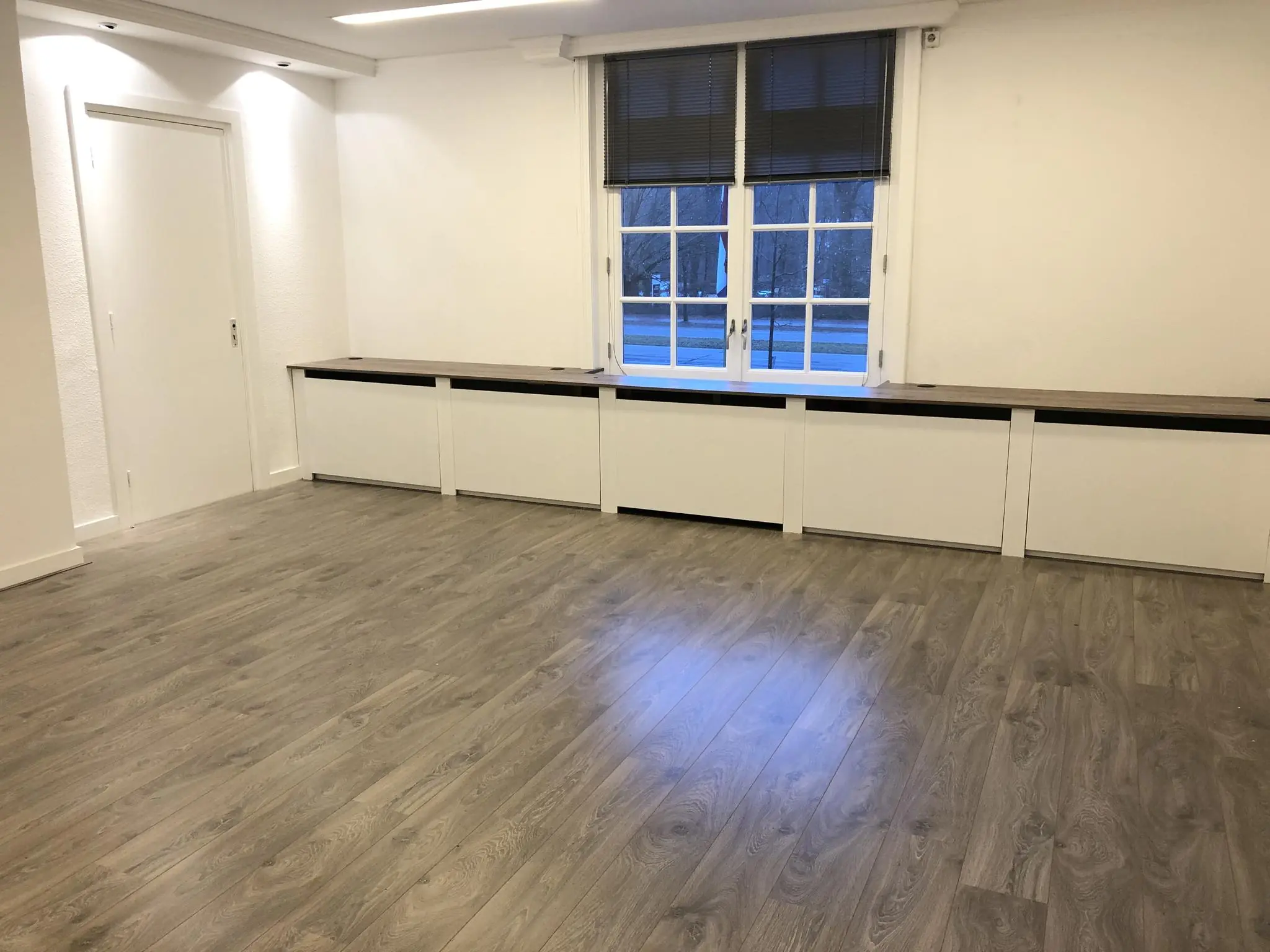 Lege kamer met houtlook vloer, witte muren, een groot raam met zwarte jaloezieën en een ingebouwde lage kast langs de muur aan de Amersfoortsestraat.