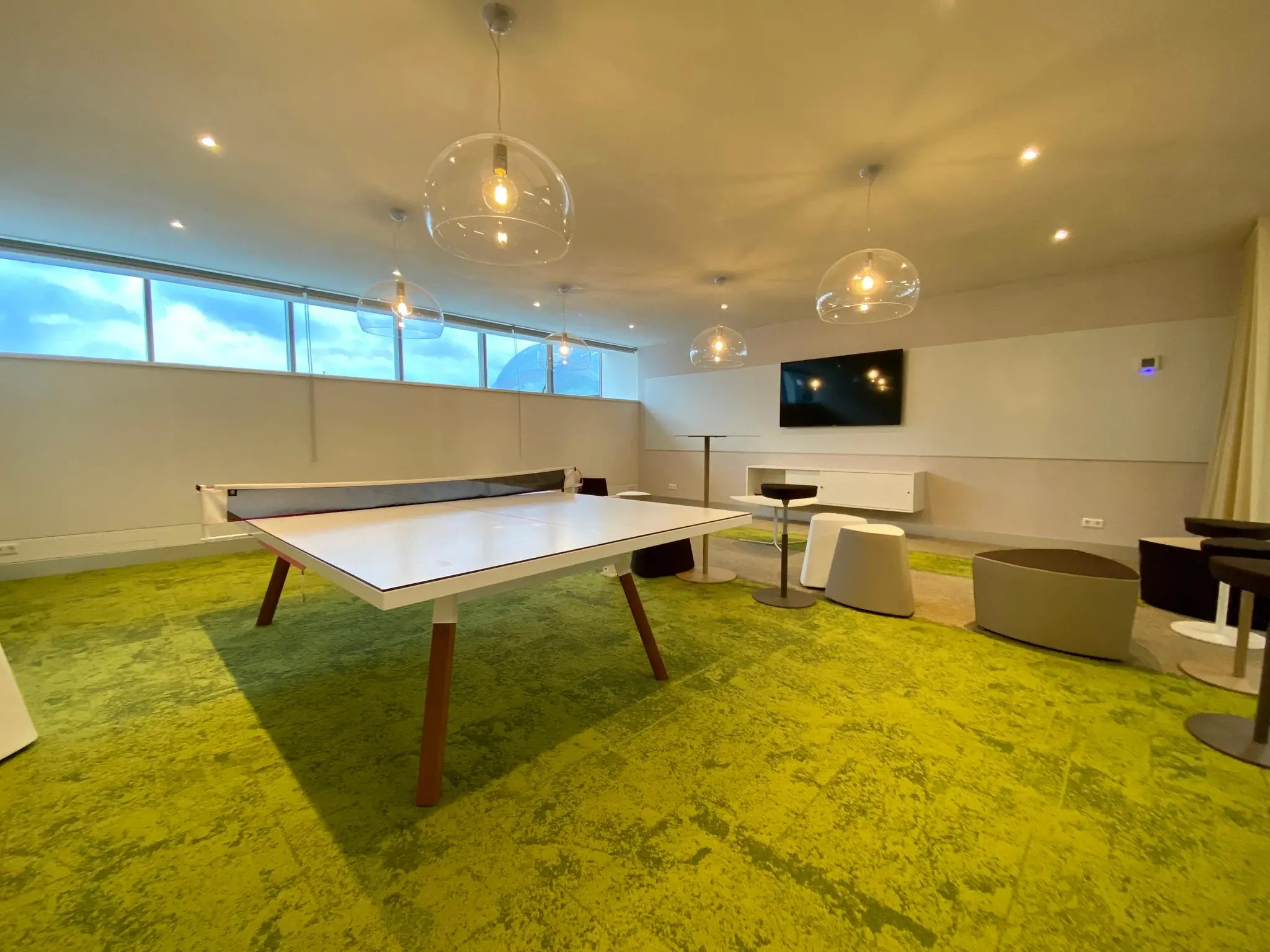Moderne recreatieruimte met een tafeltennistafel, groene vloerbedekking, zithoeken en een flatscreen-tv aan de muur.