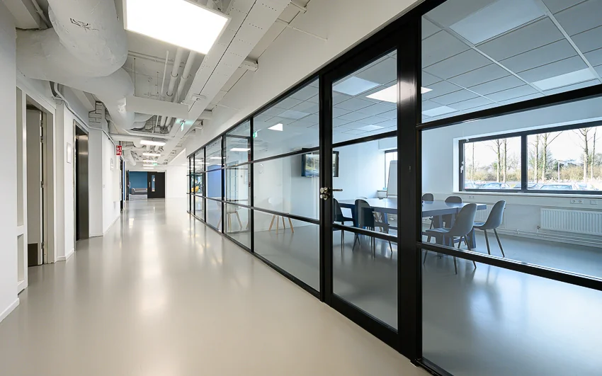 Moderne kantoorhal met glazen vergaderruimte aan de rechterzijde en zicht op buitenlicht via grote ramen.